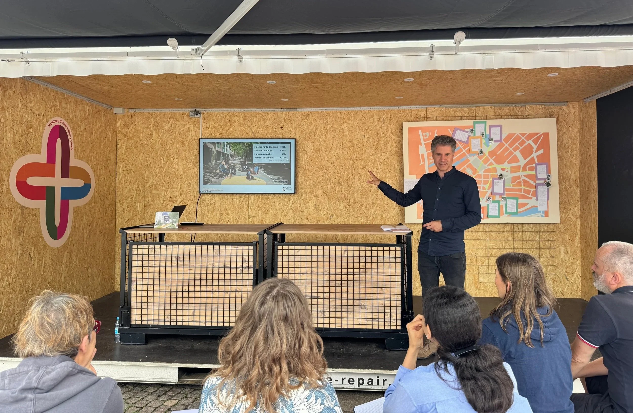 Lars Zimmermann von CITIES FOR FUTURE bei einem Vortrag im Container als Bühne. Mehrere Menschen sitzen und stehen im Publikum. Hamburg Sustainability Week 2026, Zukunftsgarten auf dem Rathausmarkt Hamburg.