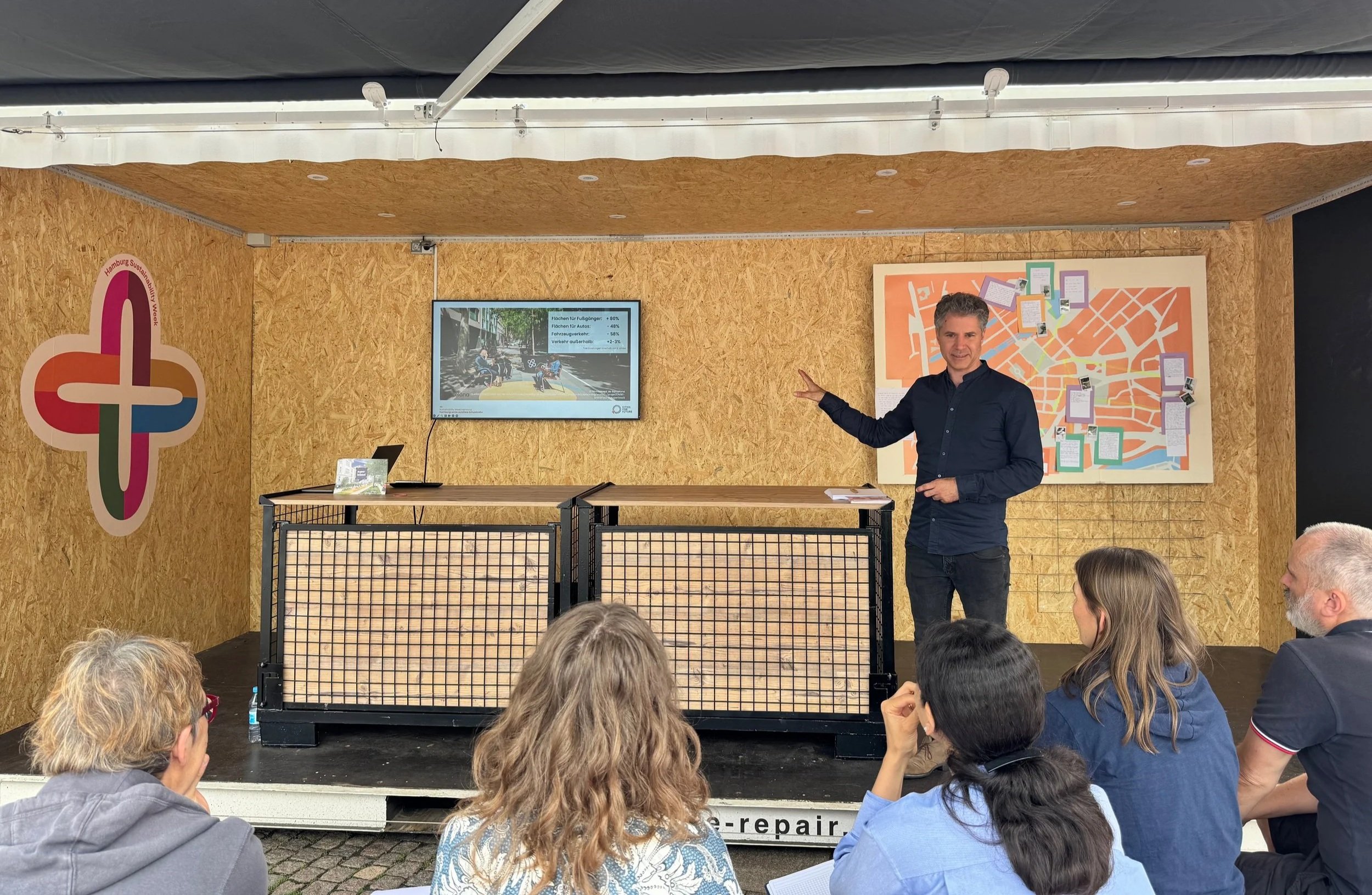 Lars Zimmermann von CITIES FOR FUTURE bei einem Vortrag im Container als Bühne. Mehrere Menschen sitzen und stehen im Publikum. Hamburg Sustainability Week 2026, Zukunftsgarten auf dem Rathausmarkt Hamburg.