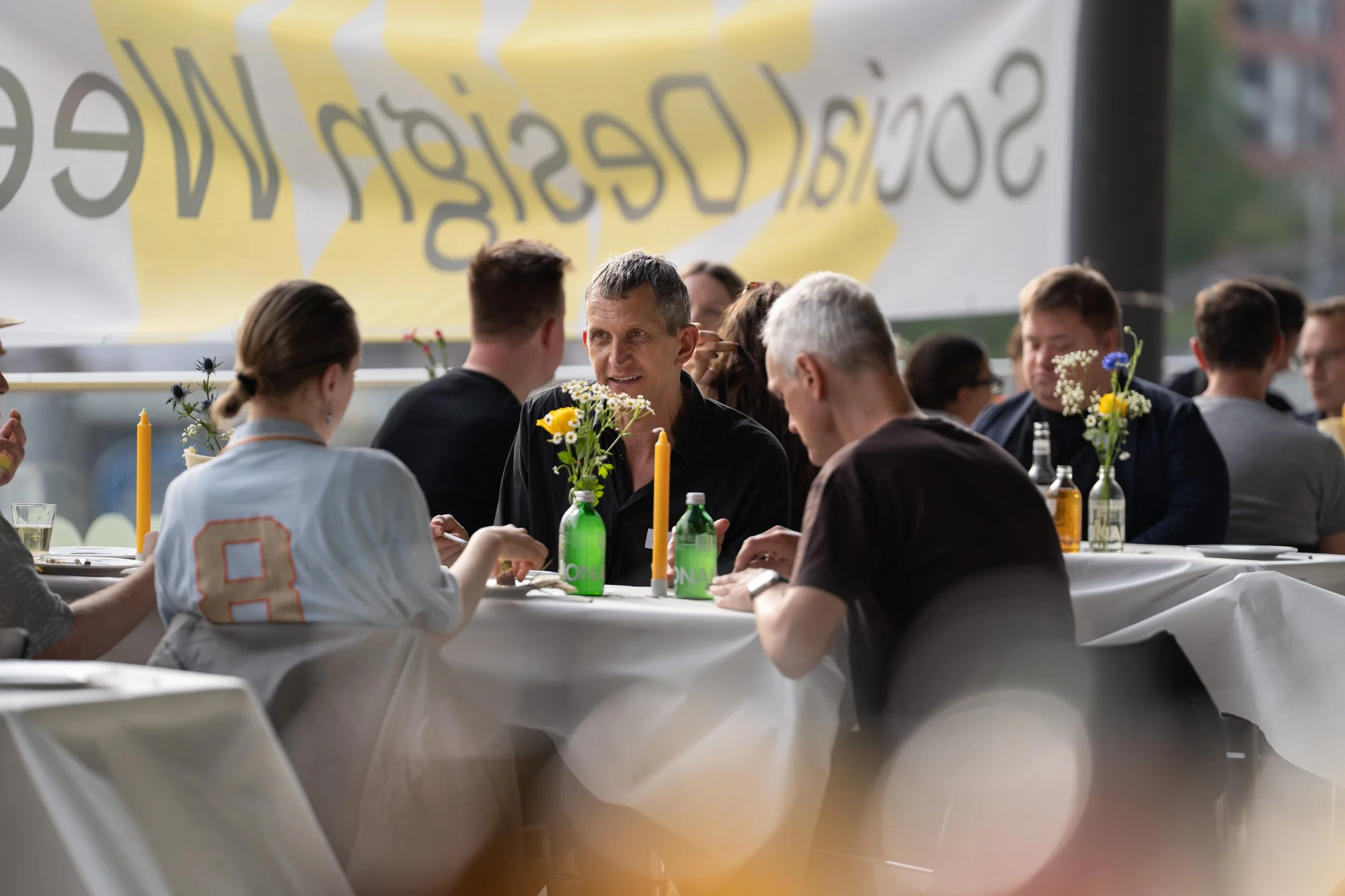 Menschen sitzen an einem festlich gedeckten Tisch mit Blumen und Kerzen, im Hintergrund ein großes Fenster mit einem gelben Banner