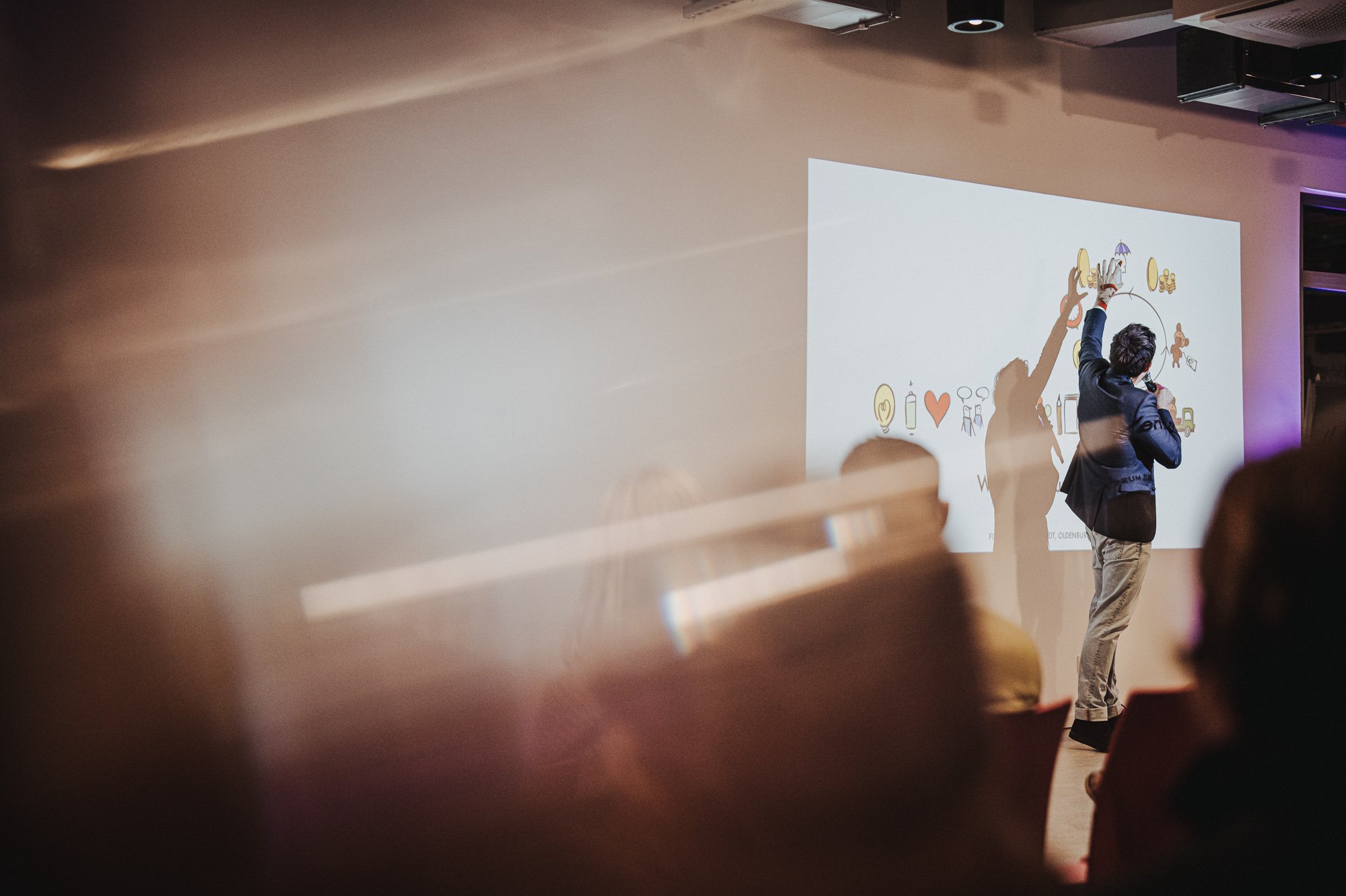 Ein Mann hält eine Präsentation vor einem Publikum. Auf der Projektionsfläche sind Symbole wie ein Herz, eine Flasche, eine Familie, und andere Illustrationen zu sehen.