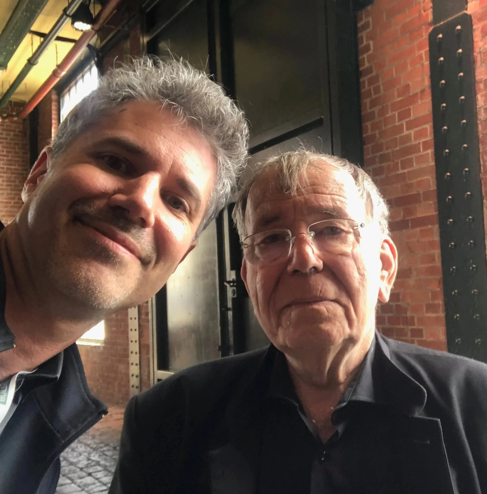 Zwei Männer, einer jünger mit grauem Haar und Lächeln, der andere älter mit Brille und grauem Haar, posieren für ein Selfie vor einer Backsteinwand im Abendlicht. Jan Gehl (Architekt aus Dänemark) und Lars Zimmermann (von CITIES FOR FUTURE)