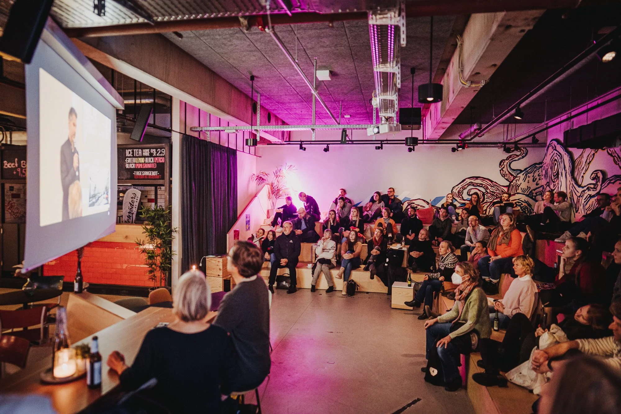 Menschen sitzen in einem abendlichen Veranstaltungsraum, sehen einen Vortrag oder eine Präsentation auf einer Leinwand, mit bunter Wandkunst und Neonlichtern.