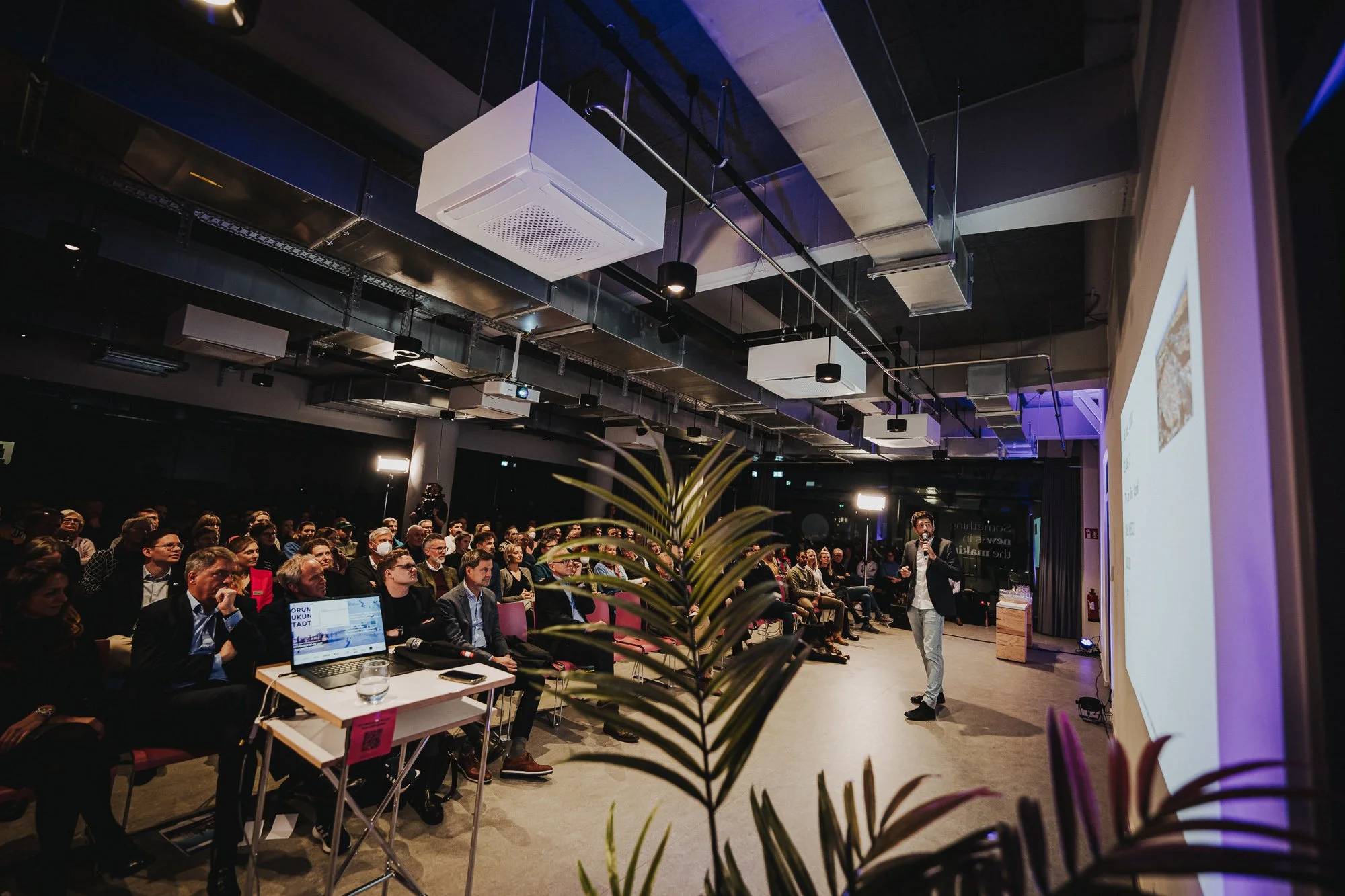 Eine Person spricht vor einem Publikum in einem dunklen Konferenzraum mit vielen Zuschauern, einer Leinwand und Pflanzen im Vordergrund.