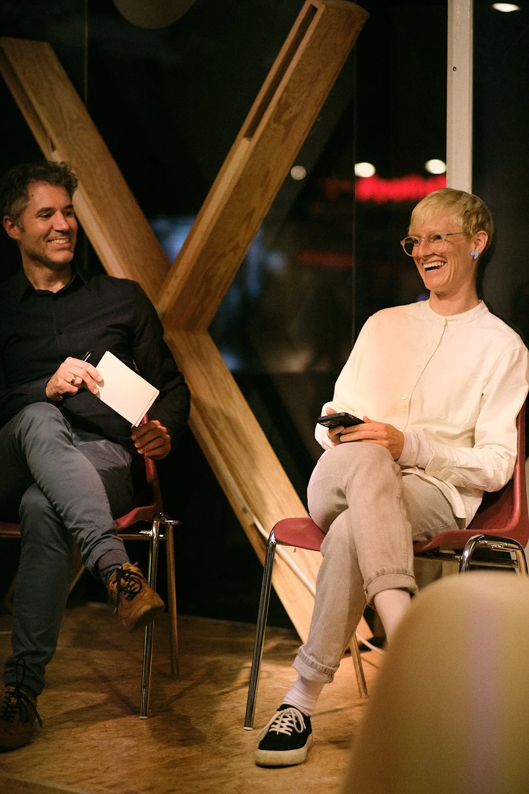 Zwei Menschen, ein Mann und eine Frau, lachen und führen ein Gespräch in einem Raum mit dunklen Wänden und Holzdekoration.