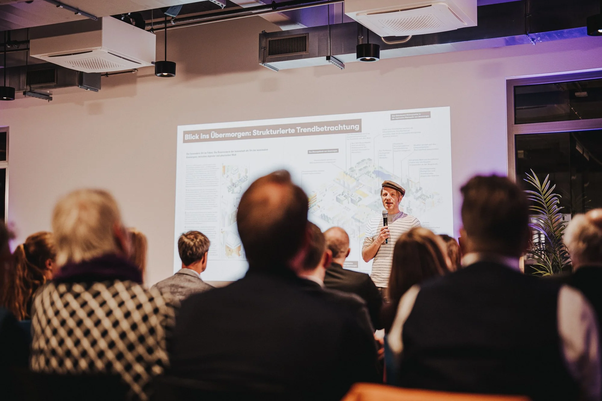 Ein Mann hält eine Präsentation vor einem Publikum in einem modernen Versammlungsraum. Er trägt ein gestreiftes Hemd und eine Mütze, spricht in ein Mikrofon und steht vor einem großen Bildschirm, der einen detaillierten Diagramm- oder Infografietitel