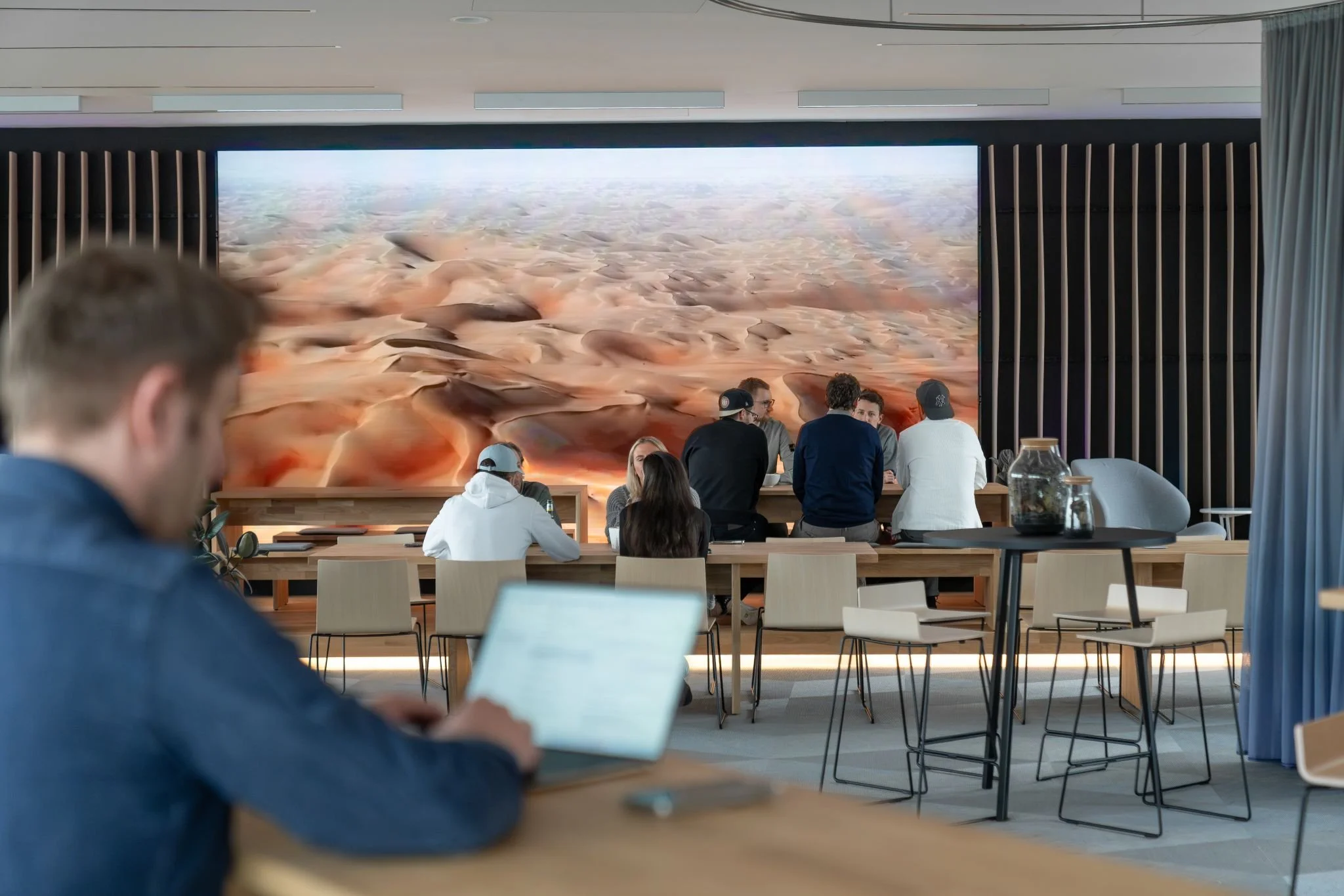 Dentsu Hamburg Büro und Cafeteria. Es ist ein Raum zu sehen wo Menschen sitzen und sich unterhalten. An der Wand ist ein Bild einer Sandwüste.