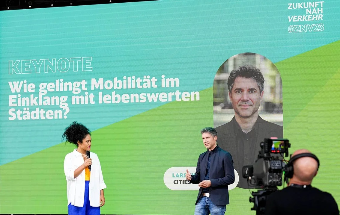 Lars Zimmermann von CITIES FOR FUTURE und eine Frau bei einer Diskussion über Mobilität in Städten. Auf dem Bildschirm steht ein Text in Deutsch: 'Wie gelingt Mobilität im Einklang mit lebenswerten Städten?'