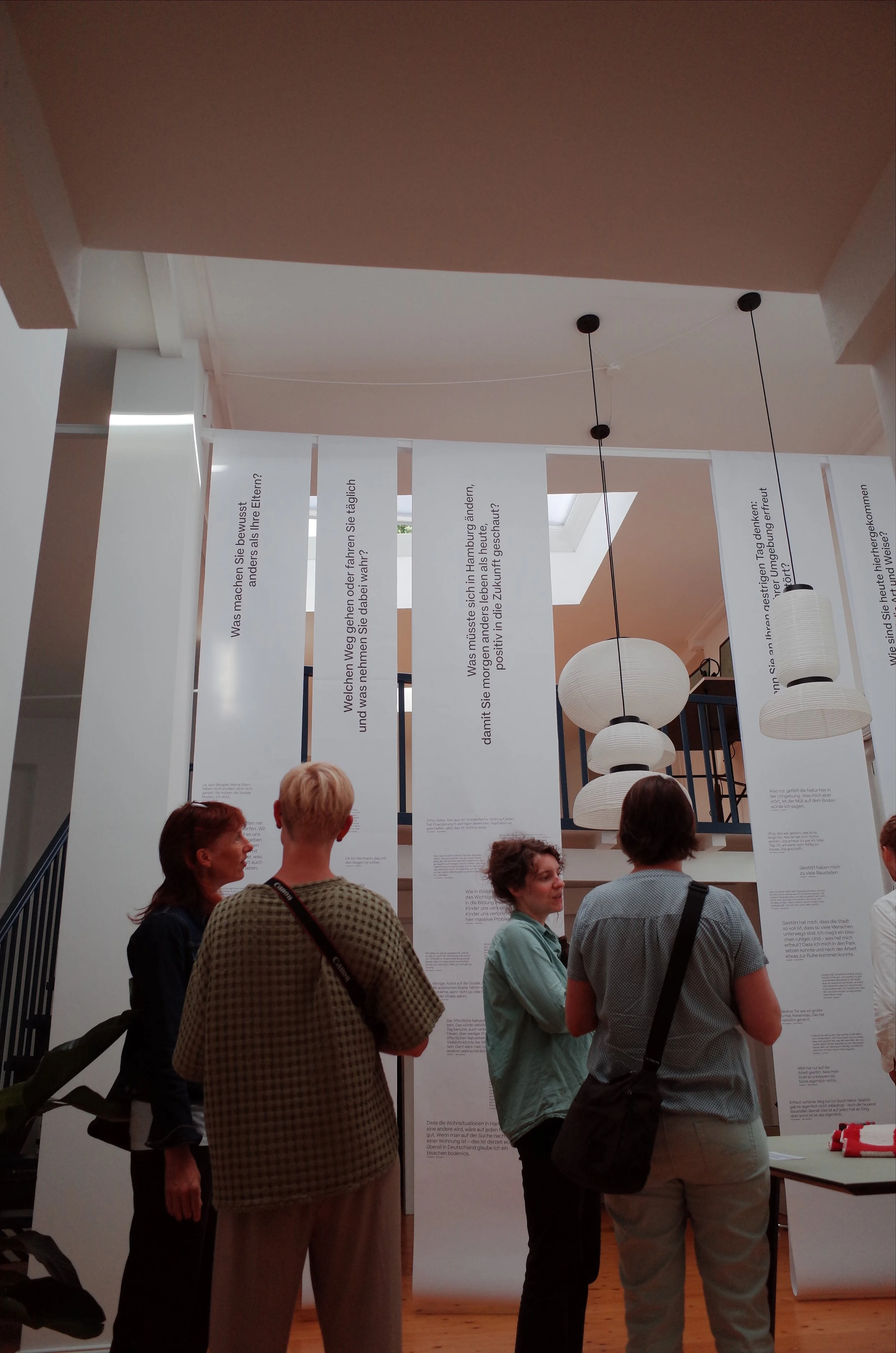 Menschen stehen in einer Ausstellung vor weißen, vertikalen Informationsflags. Es sind drei Frauen und ein Mann, die miteinander sprechen. 
Hamburg Sustainability Week 2026. Ausstellung im Loft141.
