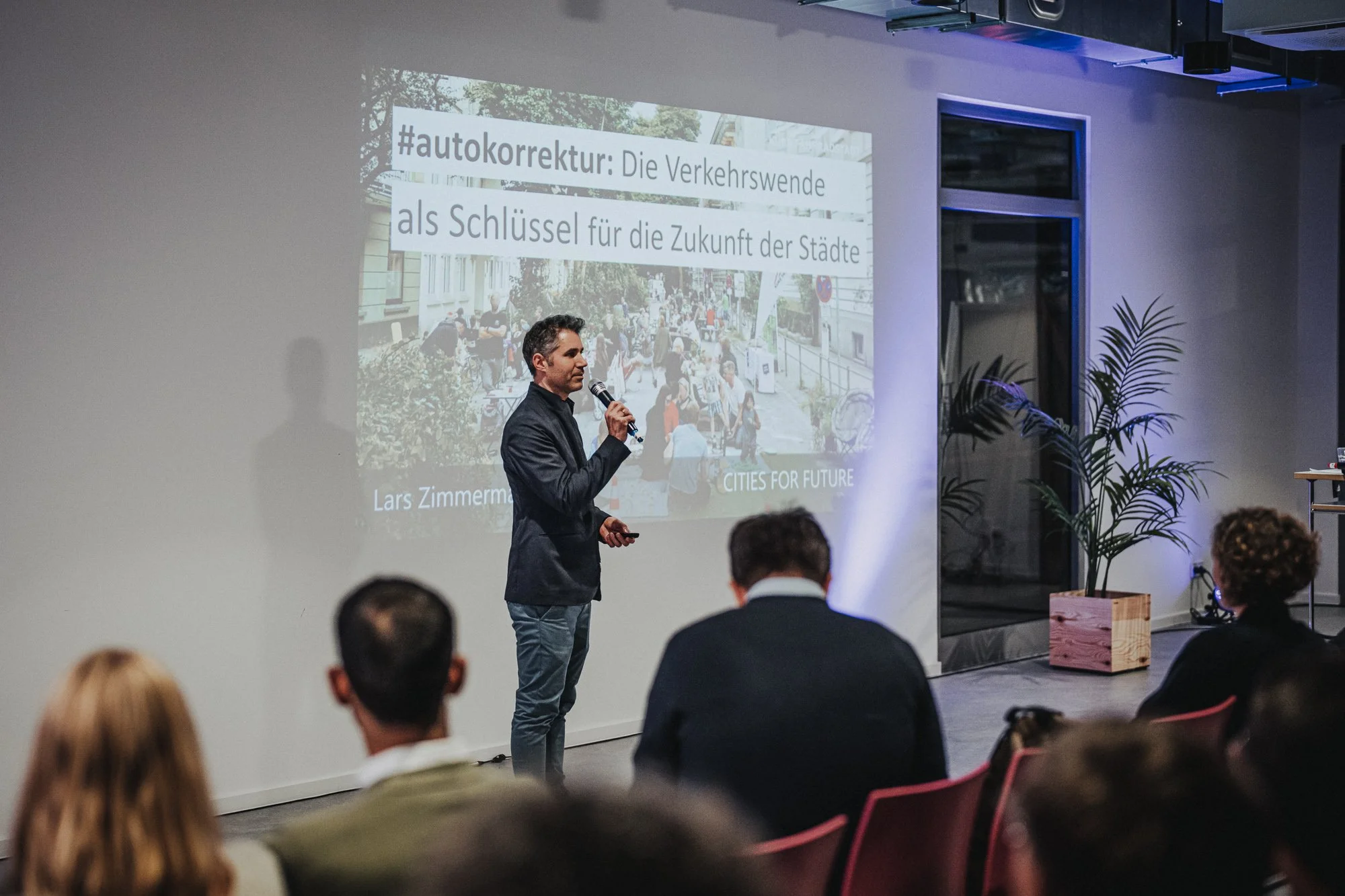 Ein Mann hält eine Präsentation vor einer Gruppe von Menschen in einem modern eingerichteten Raum. Auf der Projektionsleinwand ist ein Titel auf Deutsch sichtbar, der sich mit Verkehrswende und Stadtentwicklung beschäftigt.