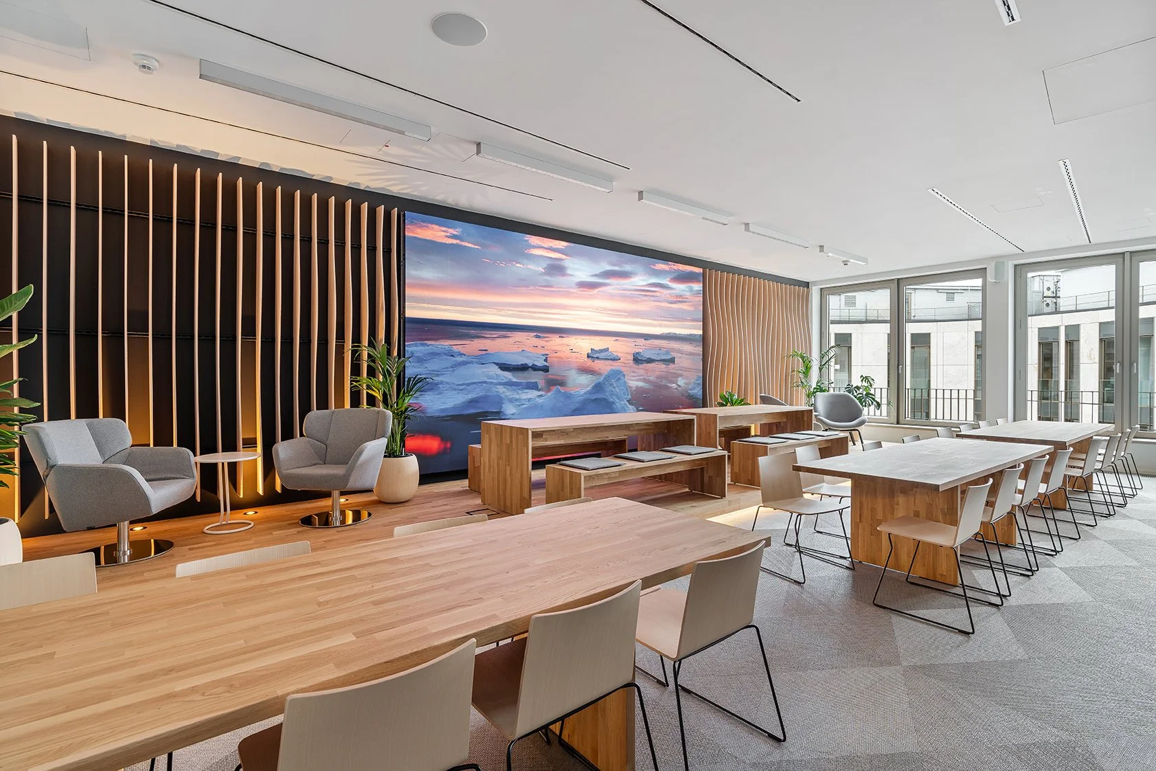 Dentsu Office: Blick in einen Raum mit langem Konferenztisch und Stühlen im Vordergrund. Hinten Sessel mit Tisch und Pflanzen vor Holzlamellenwand. Themenbereich: Interiorgestaltung, Innenarchitektur, Placemaking.