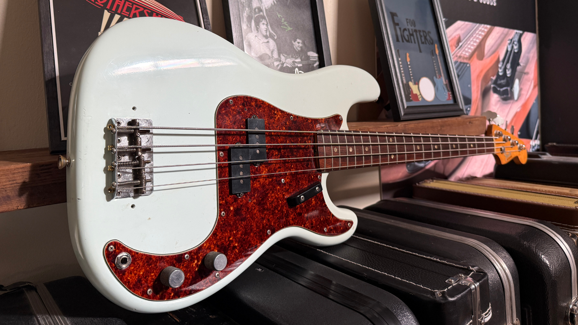 An electric bass guitar with a white body and tortoiseshell pickguard resting on a shelf among framed posters and other musical equipment.