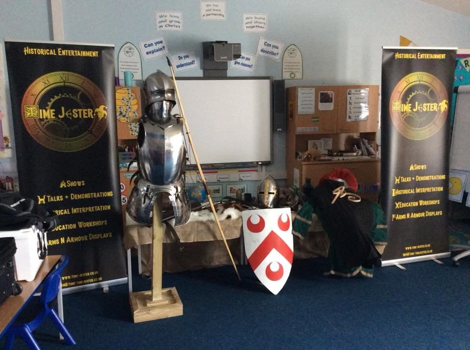 Display about historical entertainment with medieval armor, helmets, a shield with red and white crest, and banners promoting shows, talks, demonstrations, and workshops.