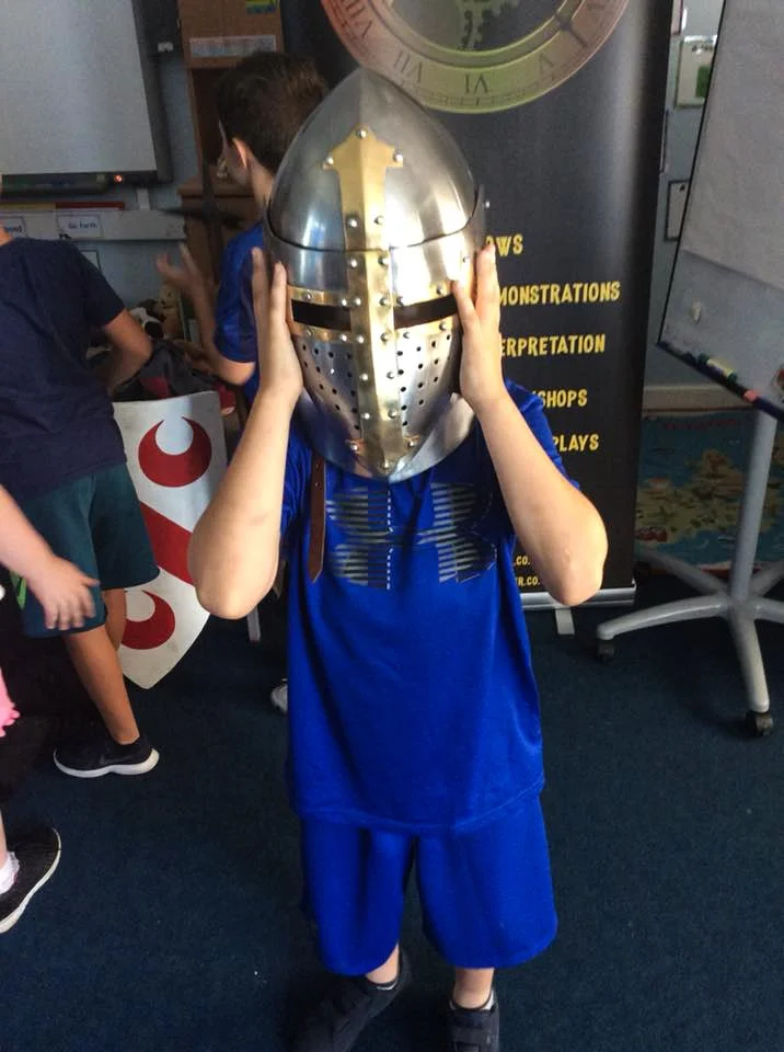 Child wearing a medieval knight helmet, holding it with both hands, in a classroom setting with other children and educational posters in the background.