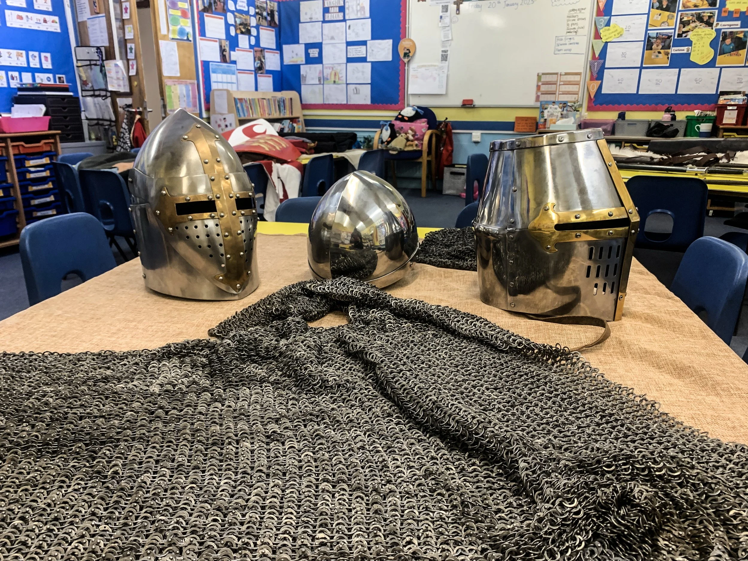 A classroom with a table displaying medieval armor helmets, a chainmail shirt, and a helmet. The background includes classroom decorations, posters, and supplies.
