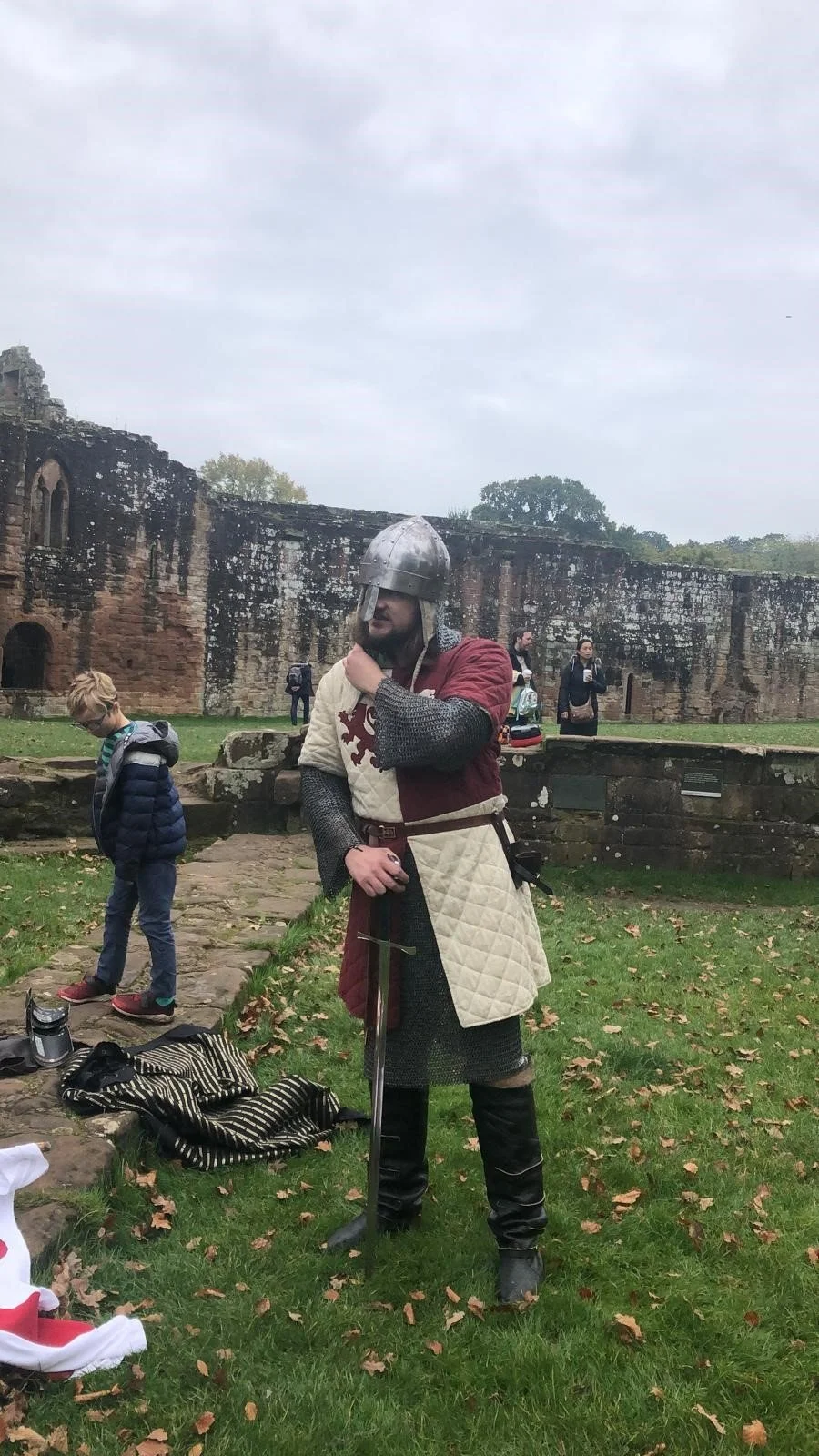 A person dressed as a medieval knight in chainmail and chainmail with a quilted tabard, helmet, and medieval sword, standing on grass near an old stone castle wall, with a few other people and children in the background.