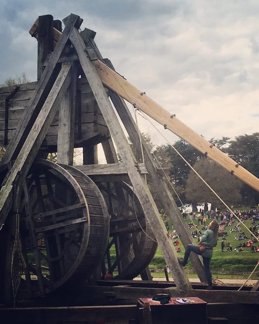 A large wooden medieval-style siege weapon, possibly a trebuchet, under construction or display outdoors with a person standing next to it. In the background, there is a hill with many people sitting and relaxing on the grass.