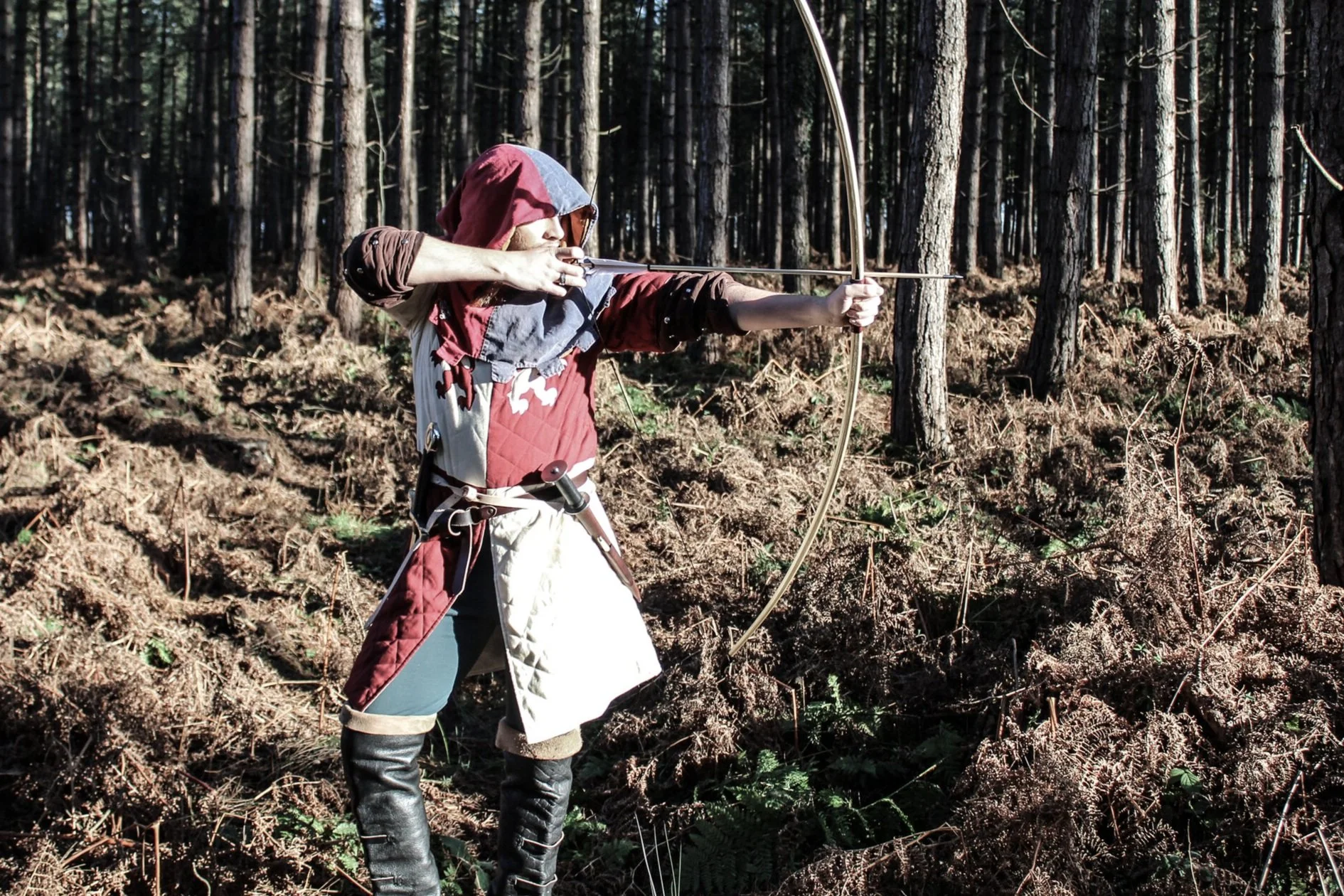 A person dressed in medieval or fantasy clothing, aiming a bow and arrow in a forest setting.