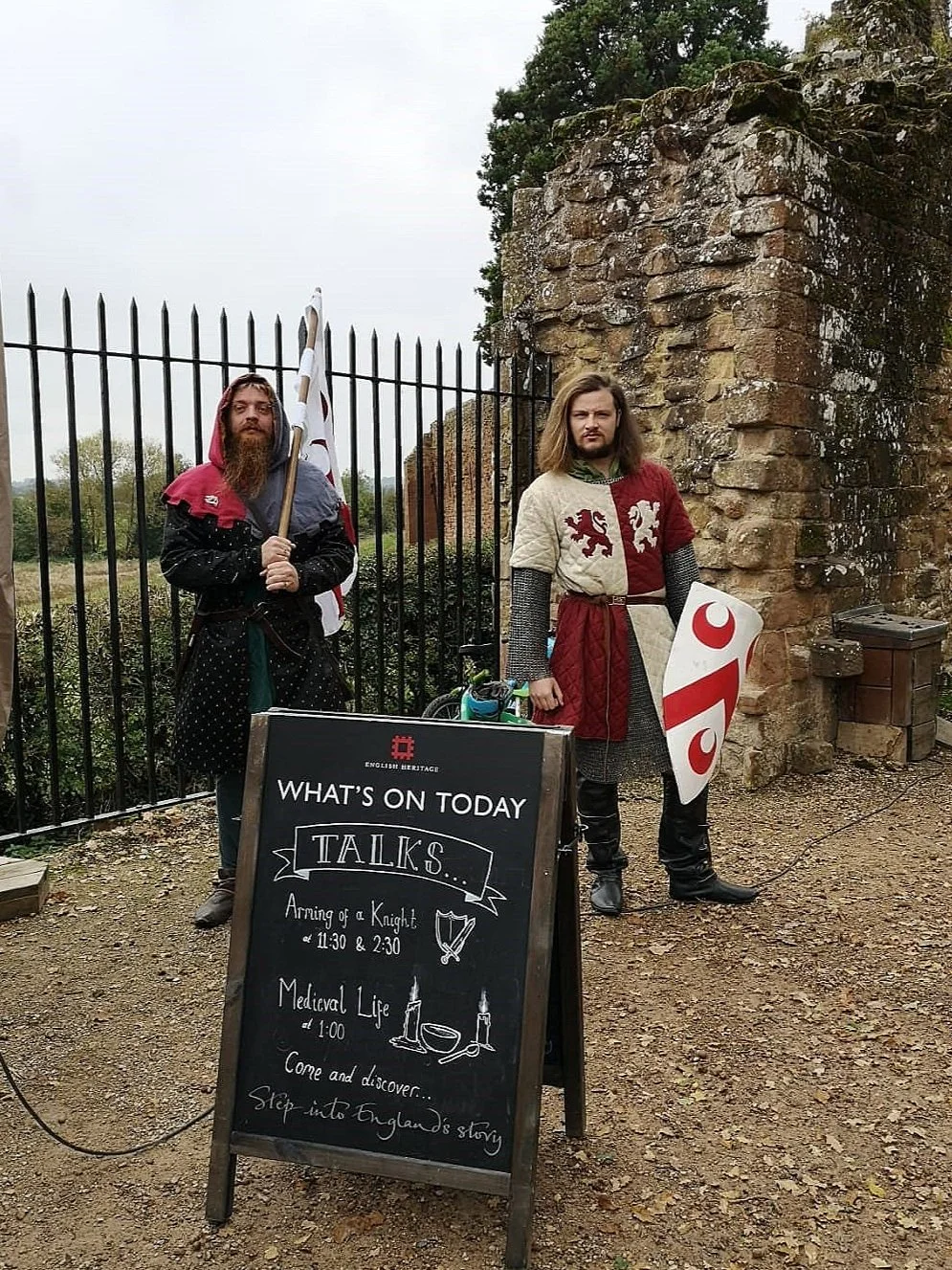 Two men dressed in medieval costumes standing outdoors near a brick structure and a black iron fence, holding shields and a flag, with a chalkboard sign advertising a medieval-themed event or talk.