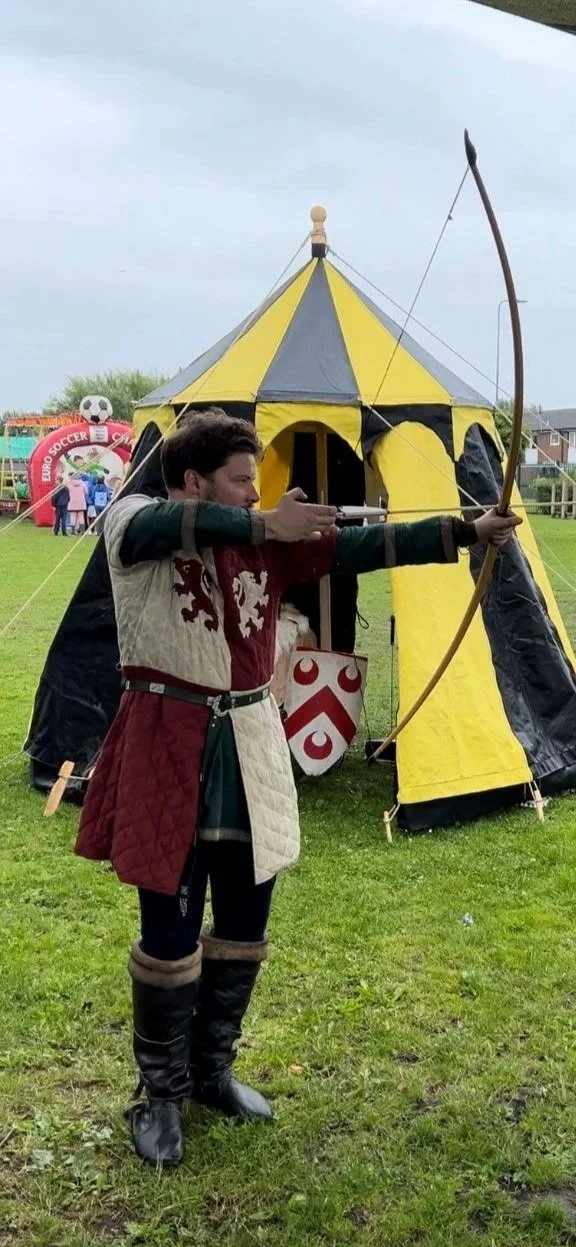 A man dressed as a medieval archer aiming a bow and arrow in front of a yellow and black tent at a fairground.