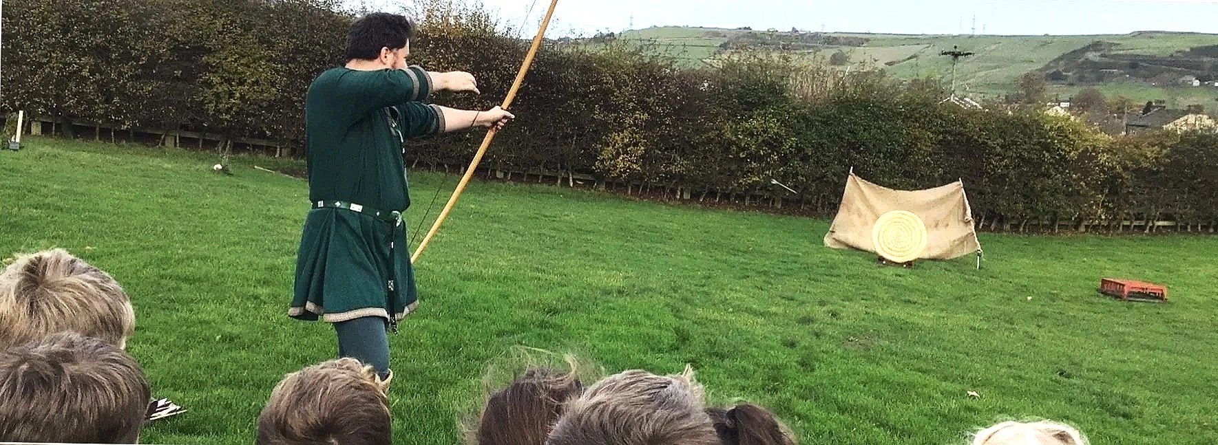 A person practicing archery outdoors, aiming a bow at a target in a grassy field with a group of children watching. The background includes bushes, hills, and a few scattered structures.