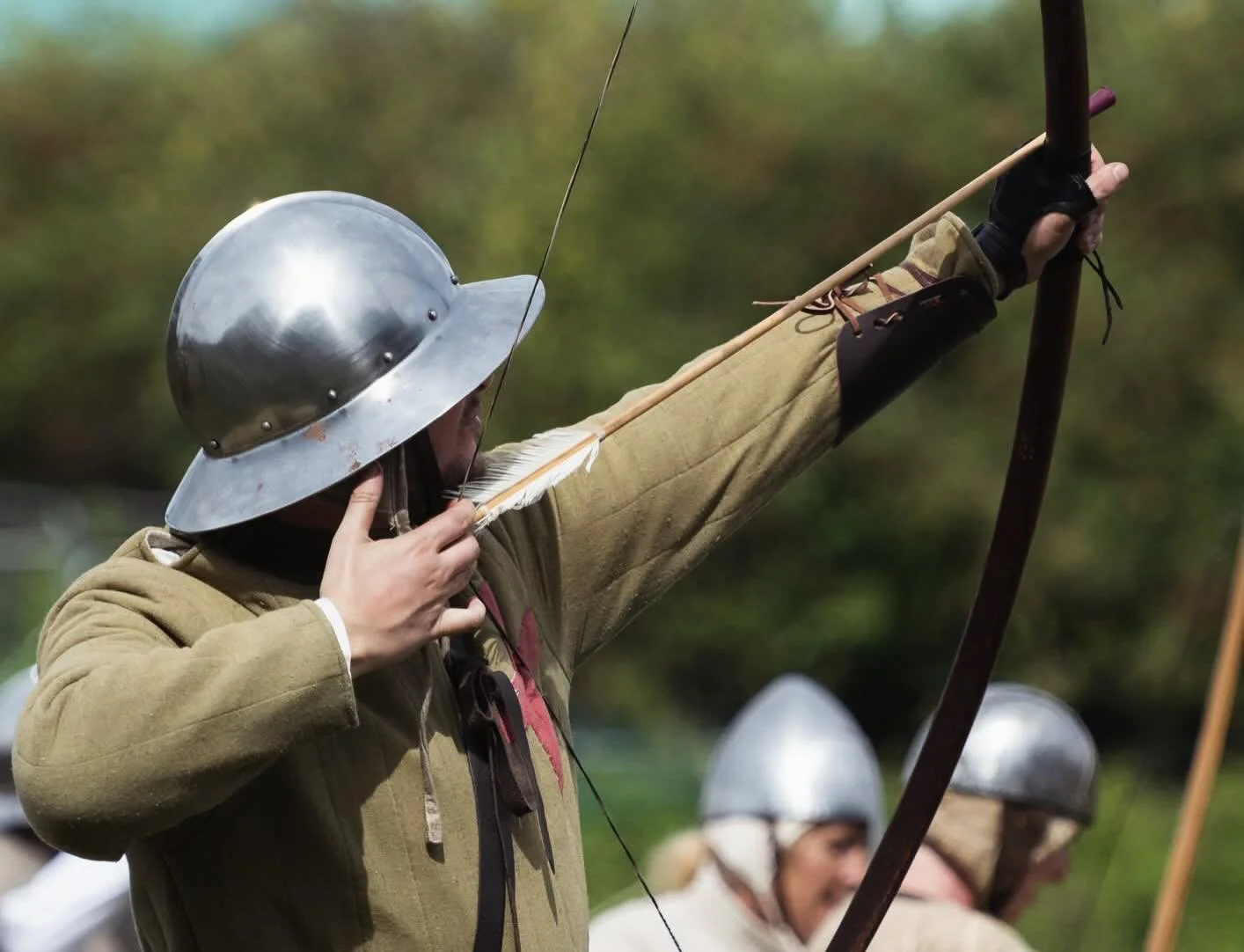Person in historical costume aiming a bow and arrow, wearing a metal helmet and beige clothing, participating in an outdoor reenactment or event.