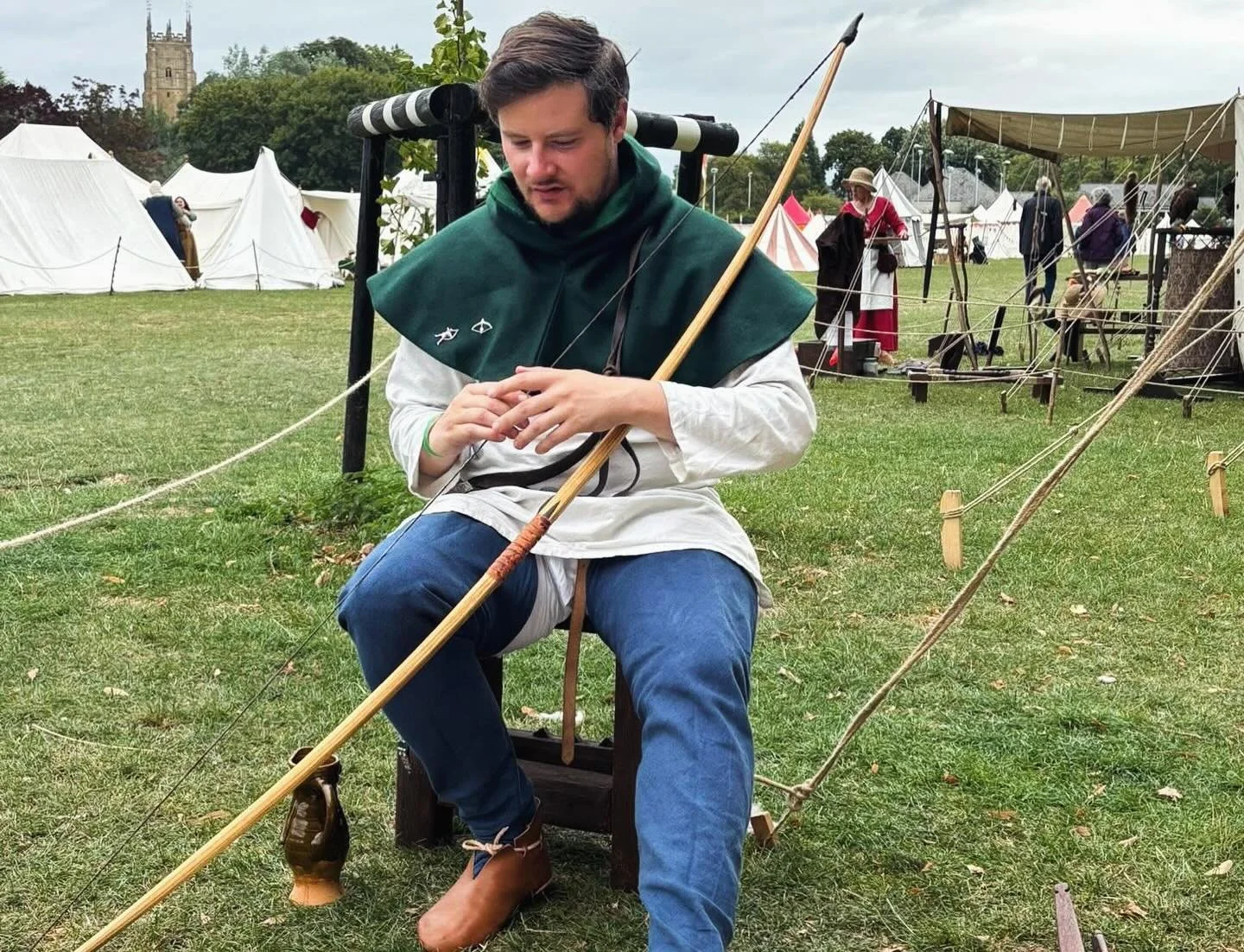 A man in medieval-style clothing sitting on a wooden stool, holding a long wooden arrow, at a historical reenactment site with tents and other people dressed in period costumes in the background.