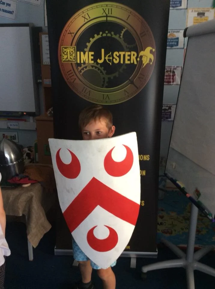 A boy standing in front of a banner that reads 'Time Jester', holding a shield with a red and white design featuring three red ovals and red chevron shapes.
