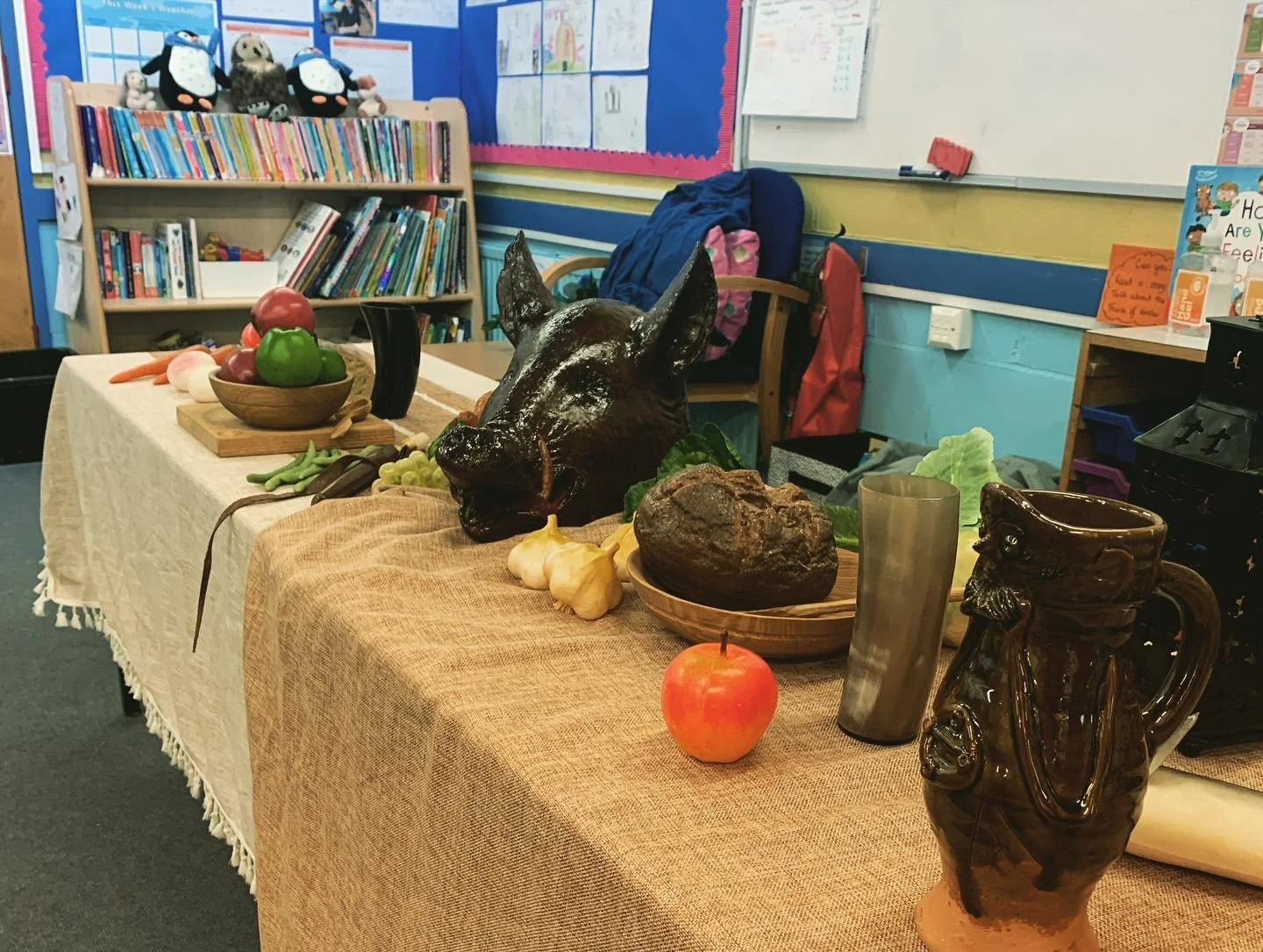 Classroom display table with a beige tablecloth holding a black wolf sculpture, carved wooden mug, apples, garlic, rocks, a glass, and various vegetables. In the background, a bookshelf with stuffed penguins, books, and educational posters are visibl