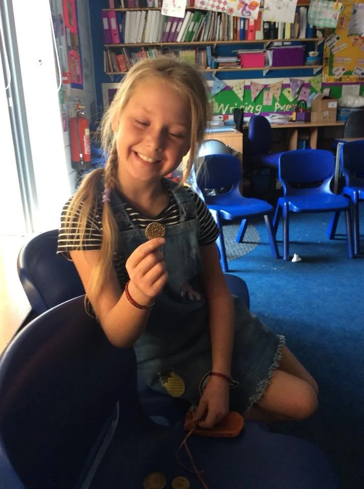 A young girl with blonde hair in a braid is sitting inside a classroom, smiling while holding a cookie. She is wearing a striped shirt and denim overalls. The background shows bookshelves, educational decorations, and blue chairs.