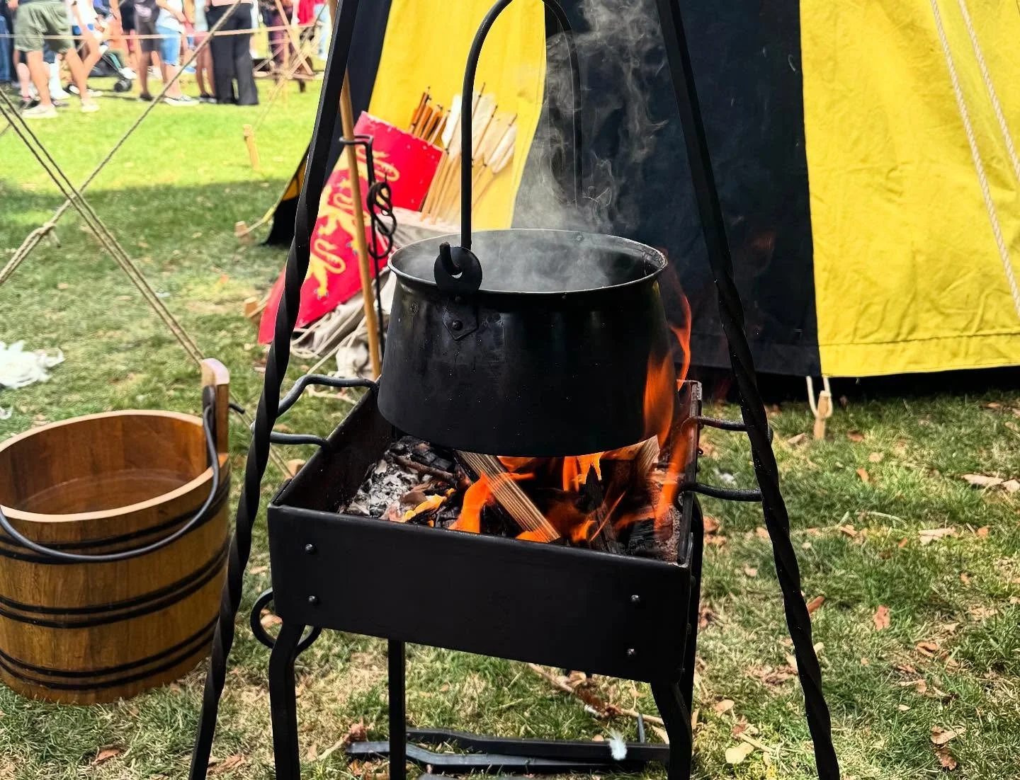 A boiling black kettle hanging over open fire on a portable stand, with a yellow tent and a crowd of people in the background at an outdoor event.