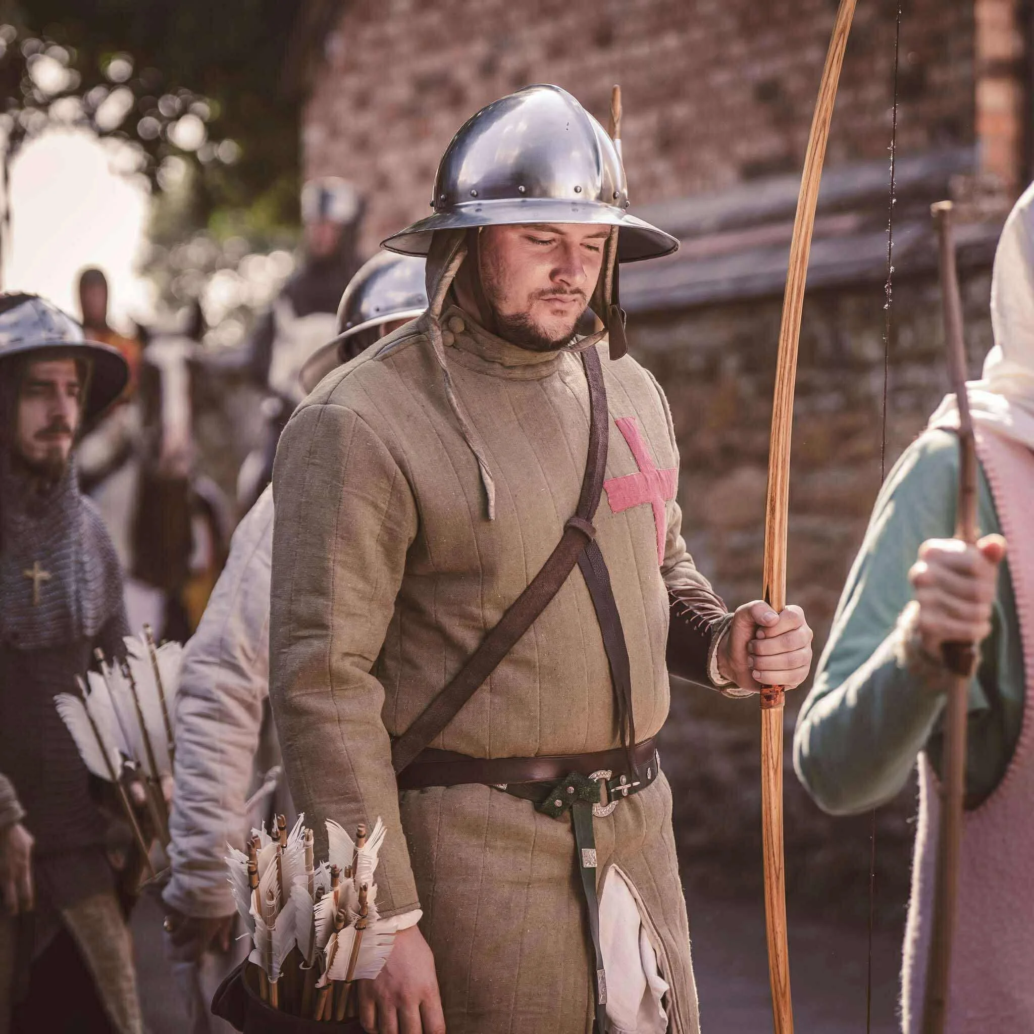 A man dressed as a medieval knight with a modern firefighter helmet, standing with eyes closed and holding a wooden staff, among other people in medieval costumes with bows and arrows, outside near a brick wall.