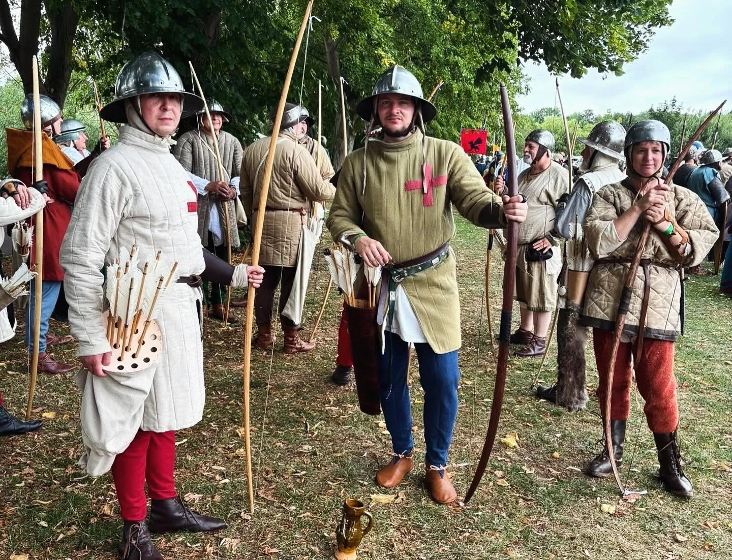 Group of people dressed as medieval soldiers or archers, wearing historical costumes and armor, holding bows and arrows, standing outdoors under trees.