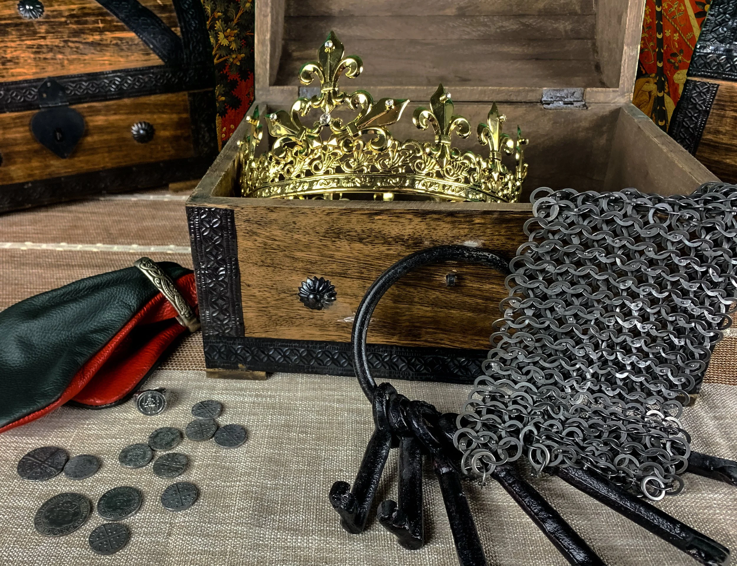 A wooden box containing a golden crown, a collection of keys, and silver chains; a pair of green and red leather gloves and scattered coins are on the beige fabric surface.