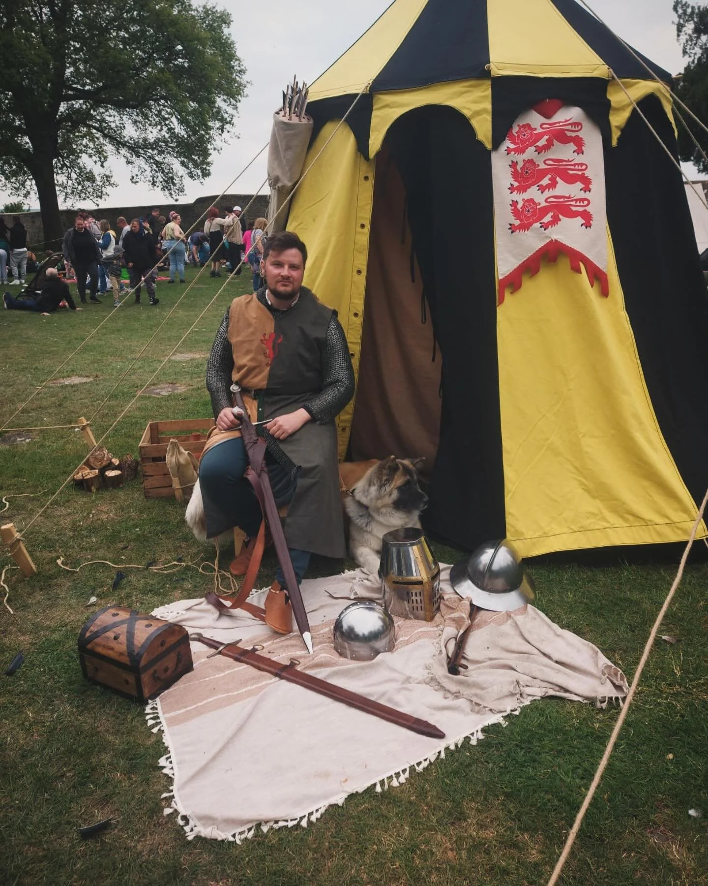A man dressed in medieval clothing poses with a dog in front of a yellow and black tent with a red lion crest. The scene includes medieval props such as helmets, a sword, and a chest, at a reenactment event with many people in the background.