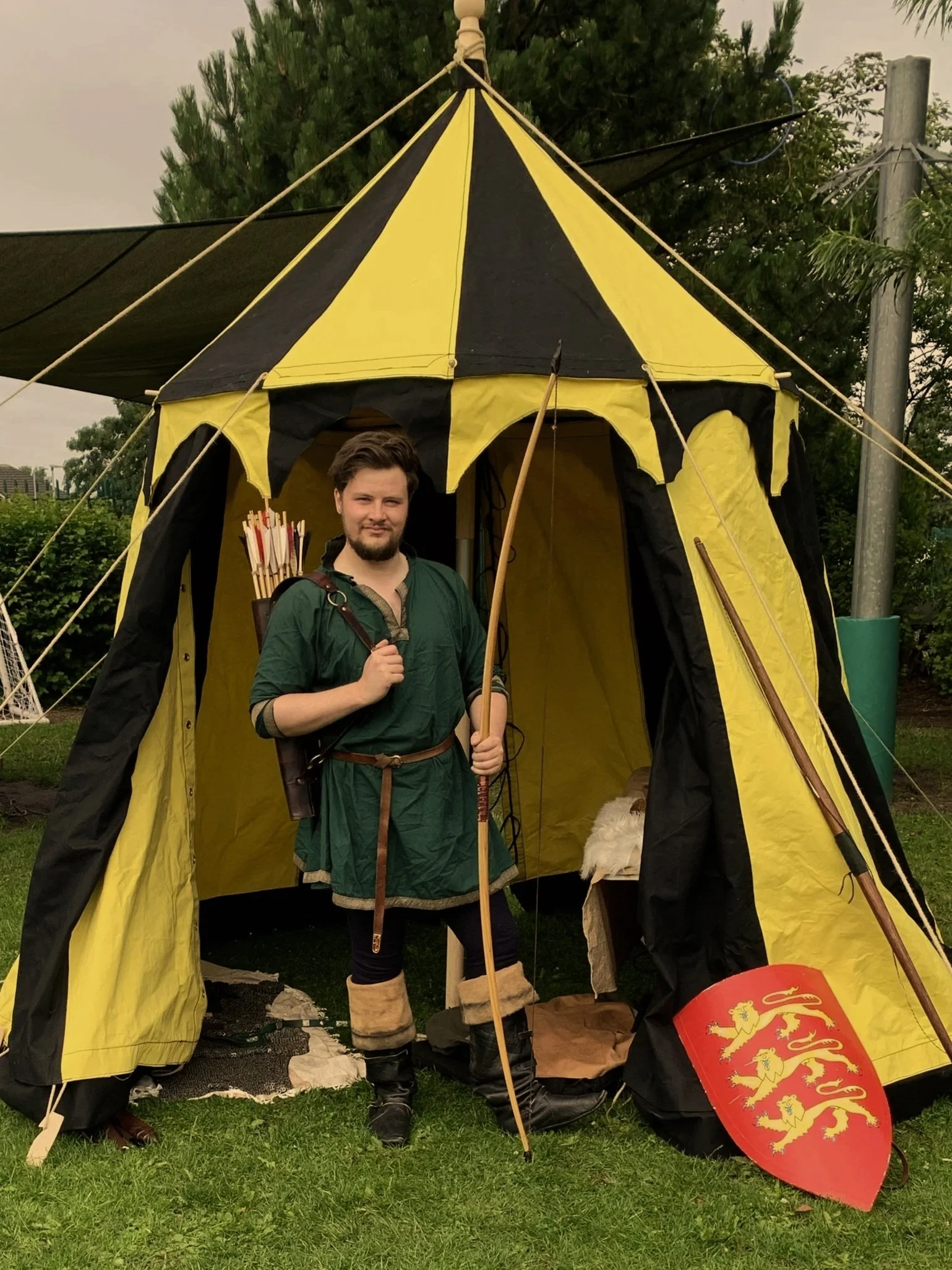 A man dressed as a medieval archer standing in front of a yellow and black tent, holding a bow, with a shield featuring three lions, outdoors on grass.