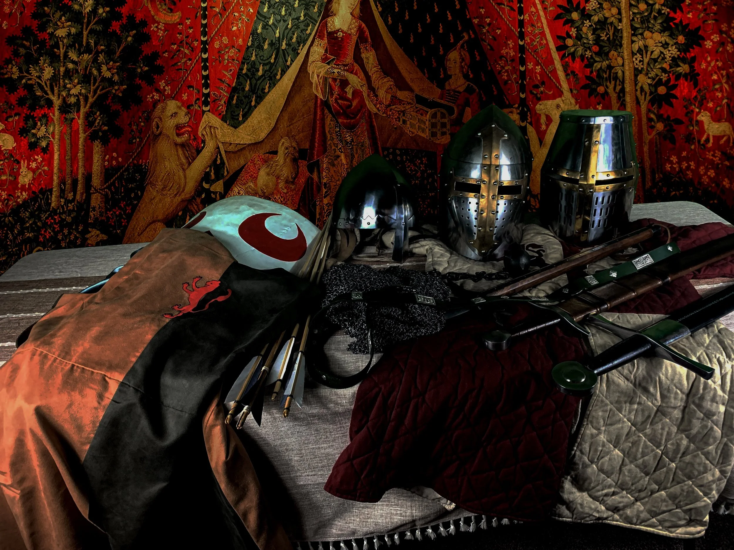Medieval knight helmets, swords, flags, and costumes arranged on a bed in front of a medieval tapestry.