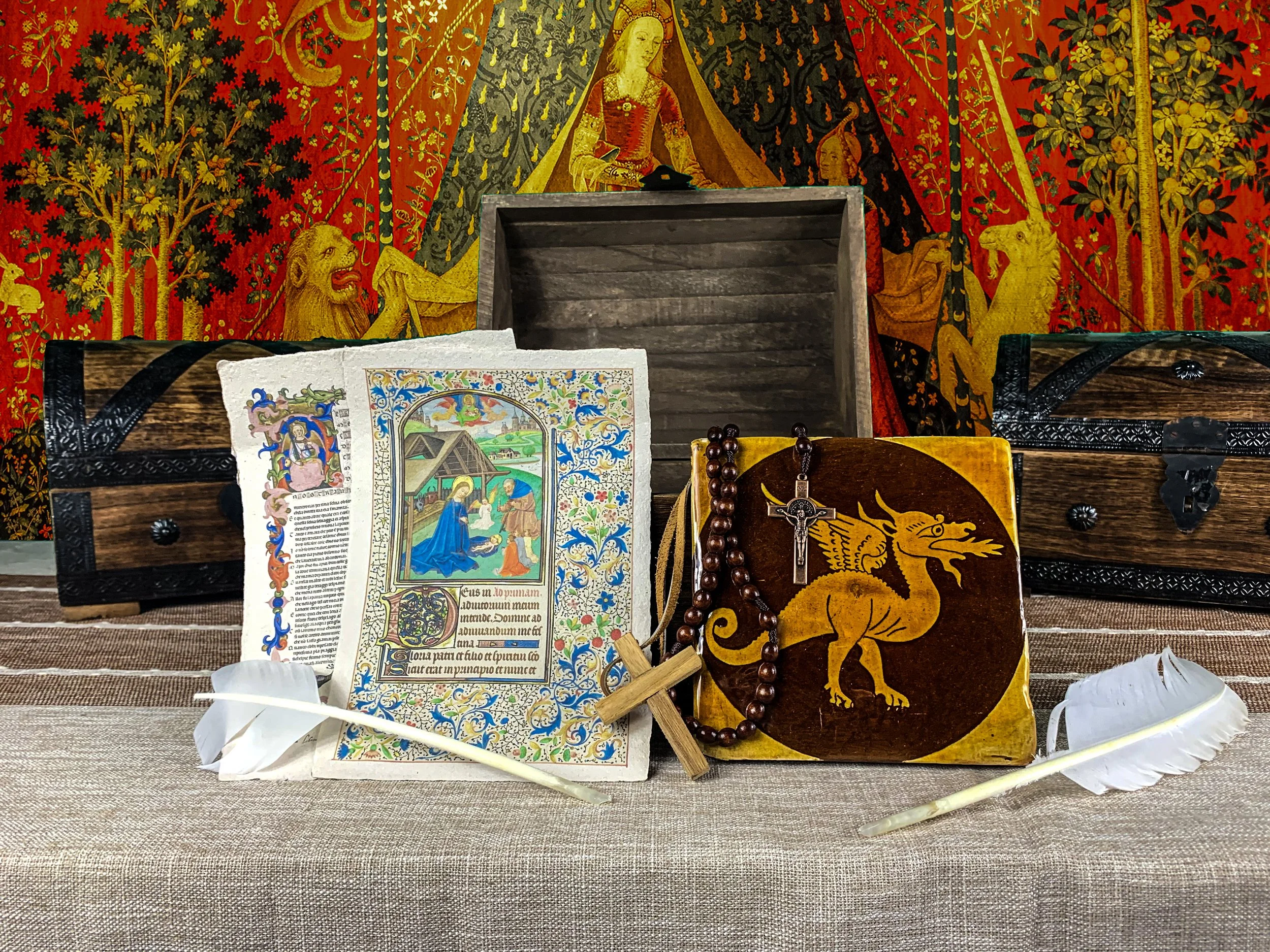 Religious artifacts including an illustrated prayer book, a wooden cross with a rosary, a leather-bound book with a dragon emblem, two white feathers, and two wooden boxes placed on a table with a tapestry of a mythological scene in the background.