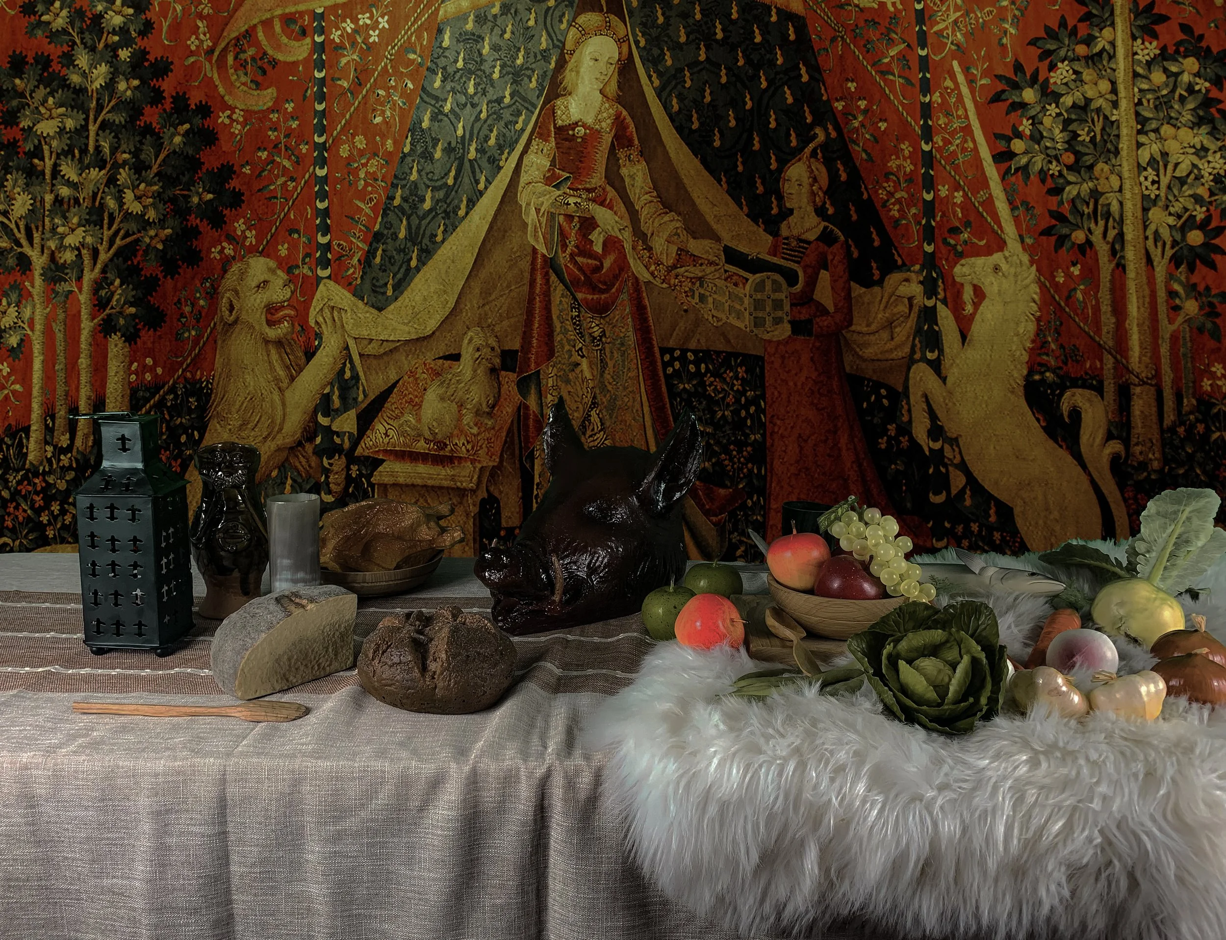 A table with a rustic cloth cover displays various food items including bread, apples, grapes, artichoke, onions, and a carved pig head sculpture. In the background, a tapestry depicting a medieval scene with a woman on a throne, lions, and a unicorn