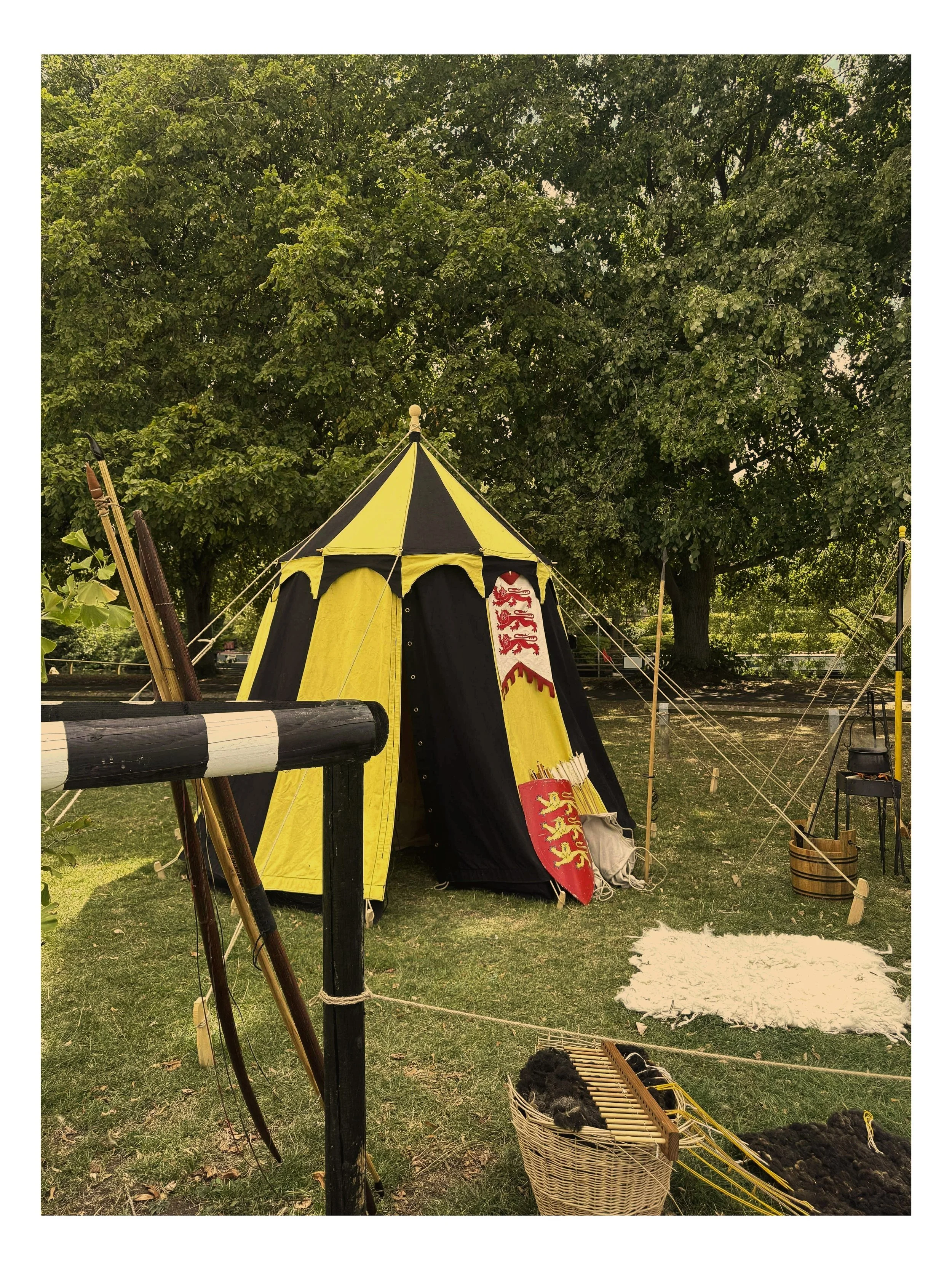 A yellow and black medieval-style tent with a crest featuring three lions at an outdoor historical reenactment setting, surrounded by trees, weapons, a small fire, and a basket of black wool or fabric.