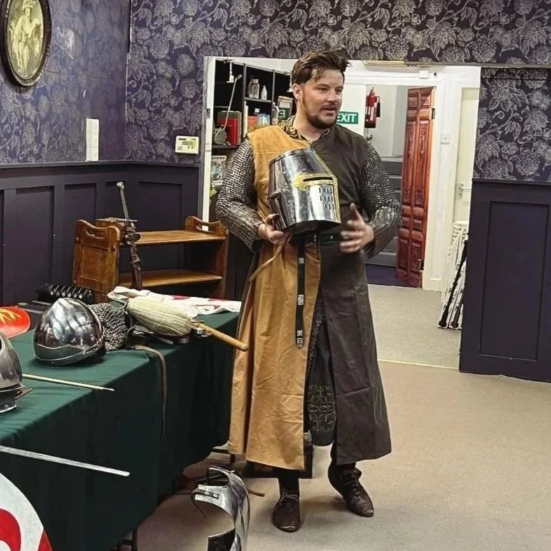 A man dressed in medieval armor, holding a piece of armor and standing beside tables displaying medieval helmets and weapons in a room with dark floral wallpaper.