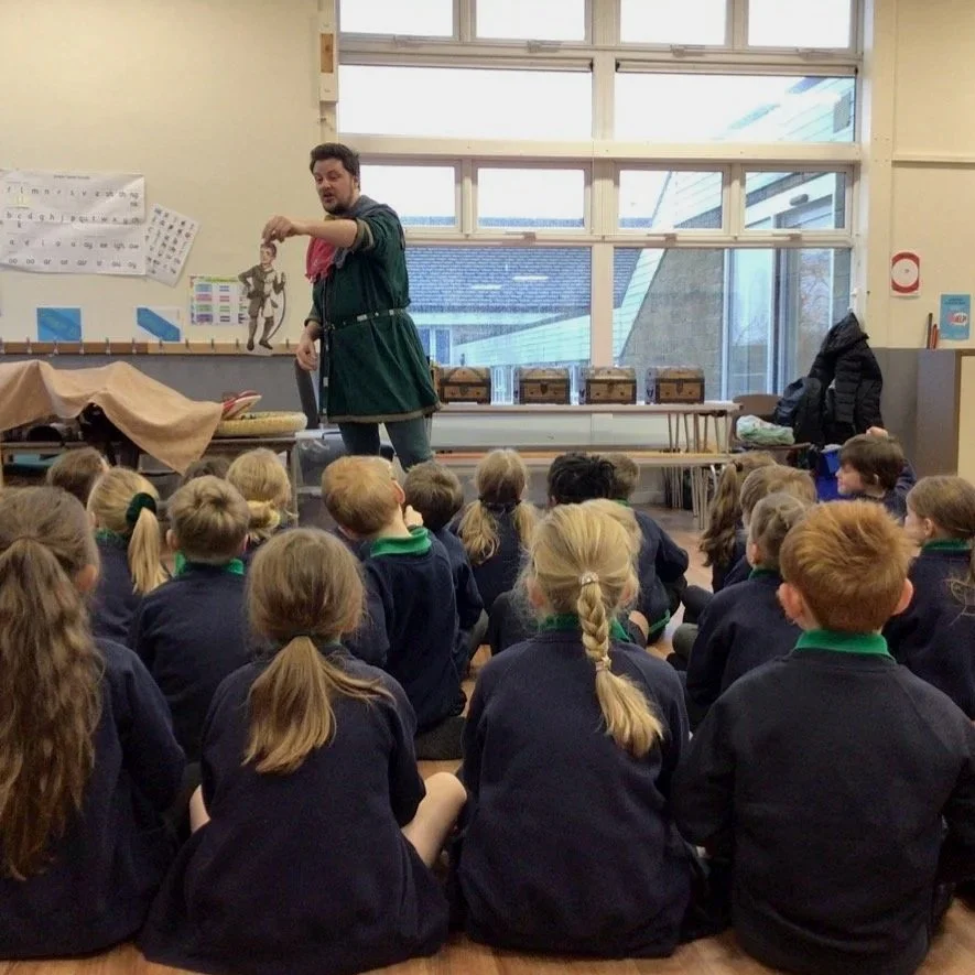 A man dressed as a medieval character in a green costume with a red sash is giving a presentation to young students seated on the floor in a classroom. The classroom has large windows, posters, and boxes in the background.