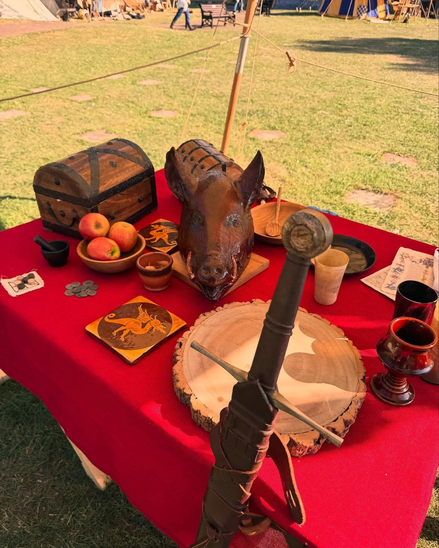 A table covered with red cloth displaying medieval-themed items including a roasted pig's head, a wooden treasure chest, apples, coins, candles, and decorative cups, set outdoors on grass with tents and people in the background.