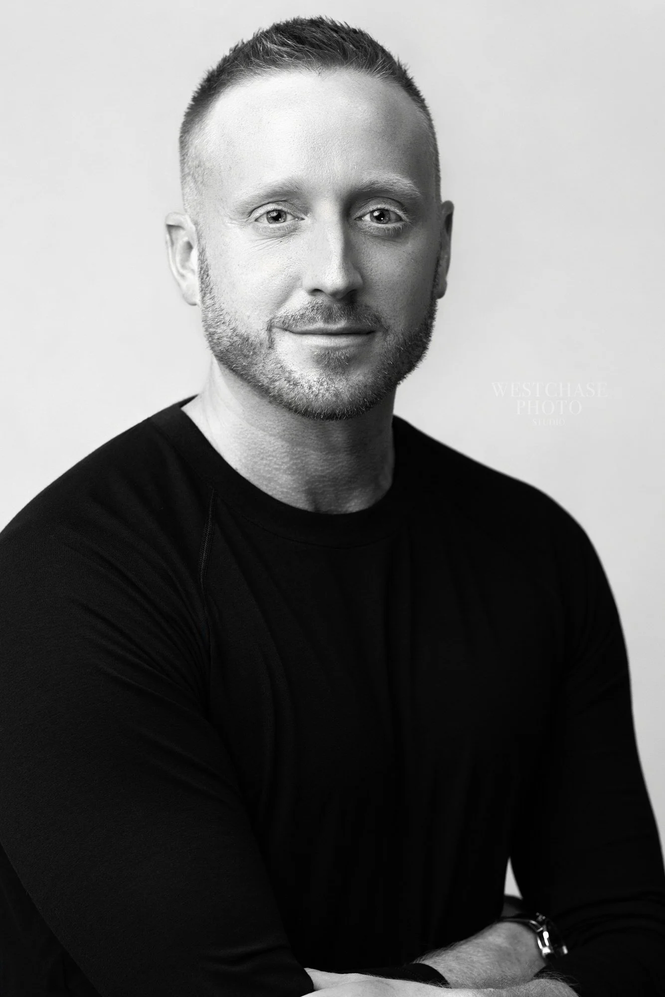 Black and white portrait of a man with short hair and a beard, smiling slightly, wearing a long-sleeved black shirt, sitting with arms crossed, in front of a plain background.