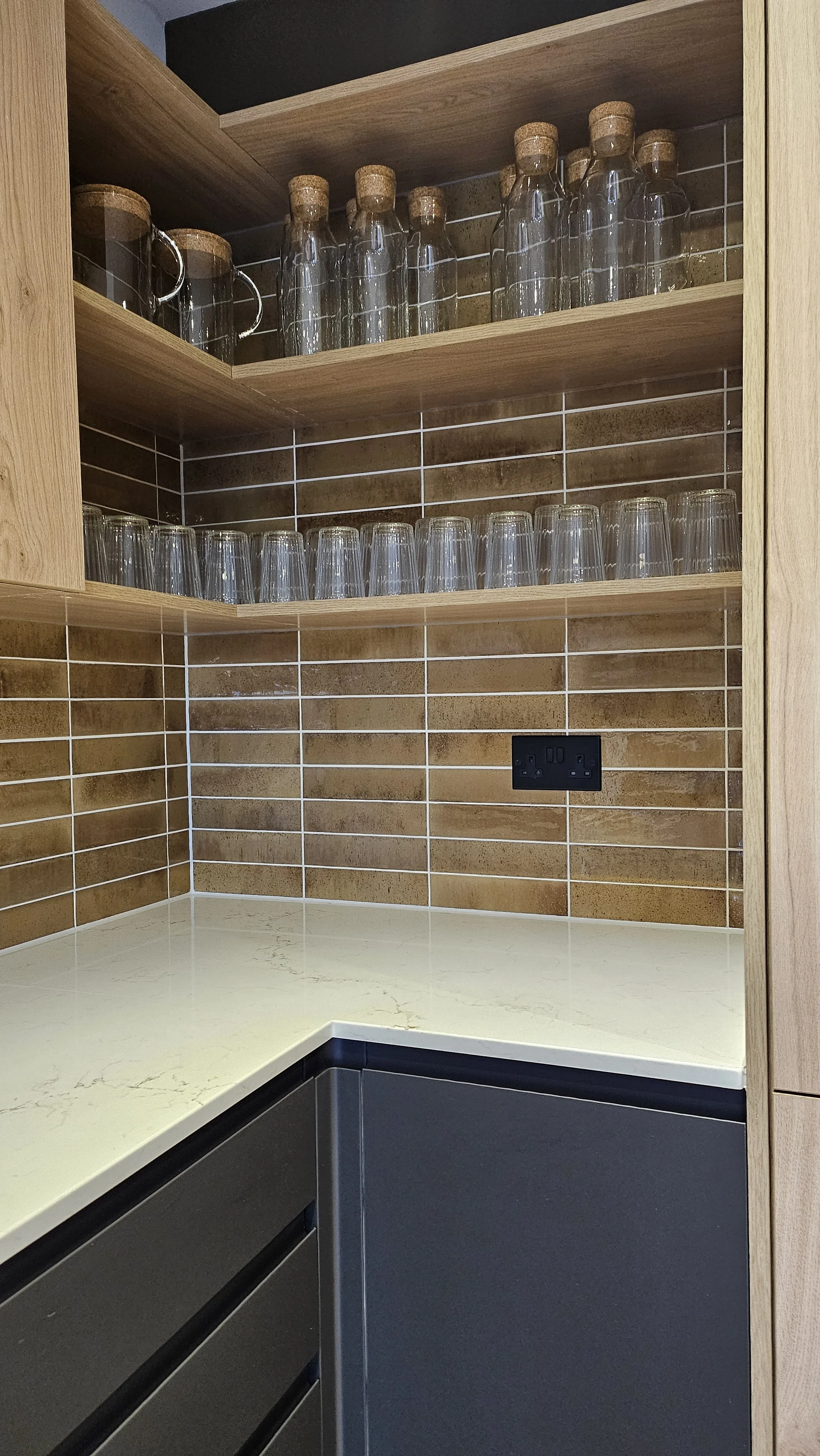 Kitchen corner with beige marble countertop, brown tiled backsplash, wooden open shelves with glass bottles and cups, and a black electrical outlet.