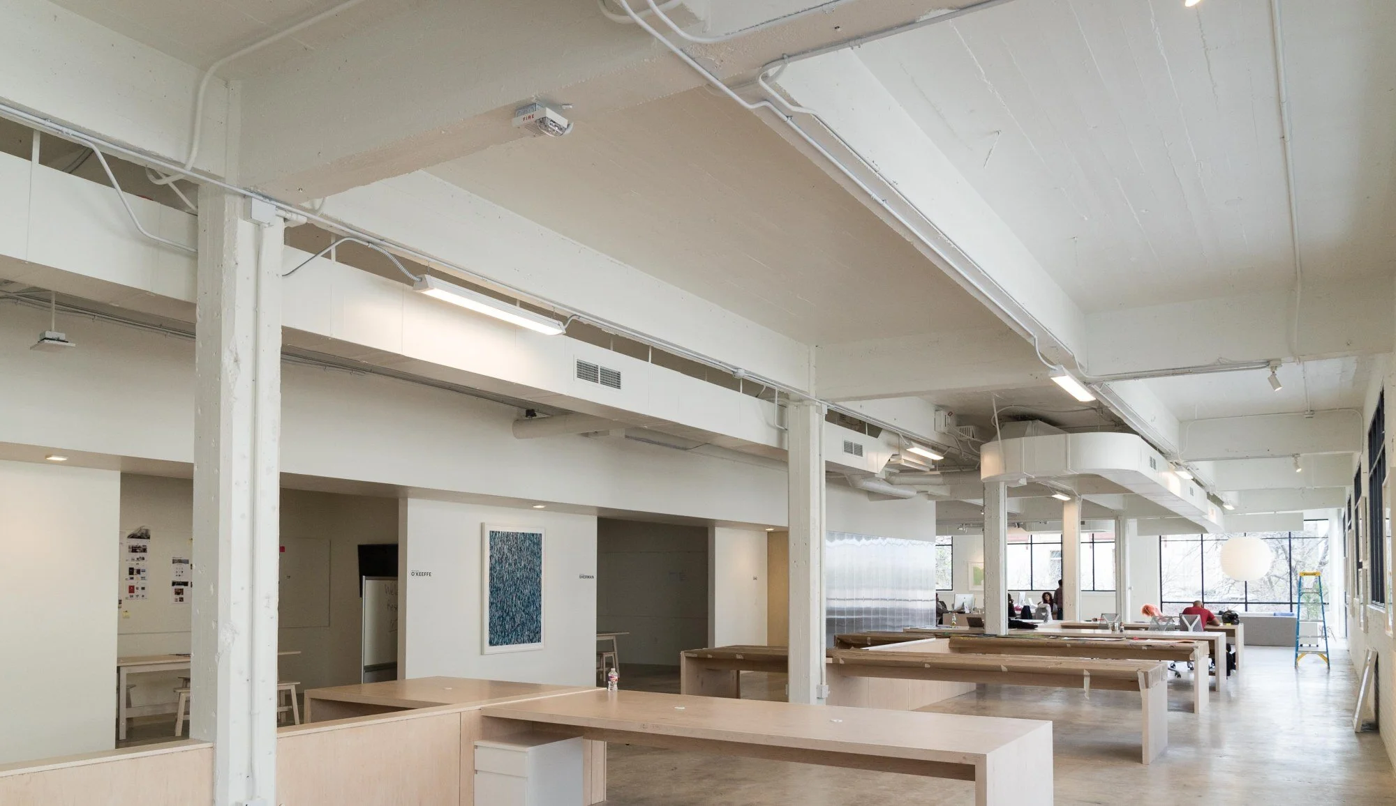 Spacious, modern, and well-lit interior office space with minimalist furniture, large windows, and a few people working in the background.