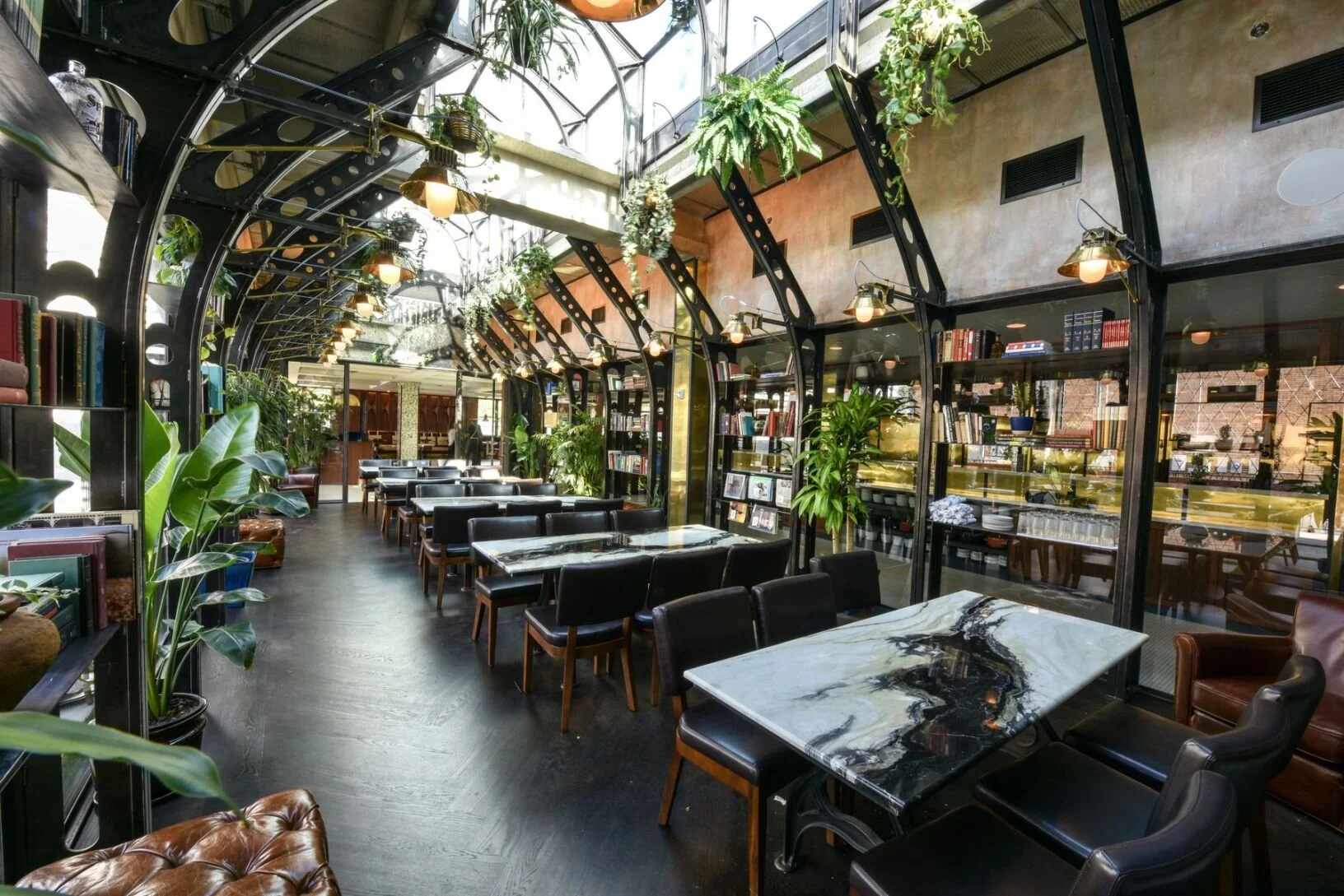 Elegant restaurant interior with black leather chairs, white and black marble tables, and large green plants. Glass ceiling allows natural light, complemented by warm lighting fixtures.
