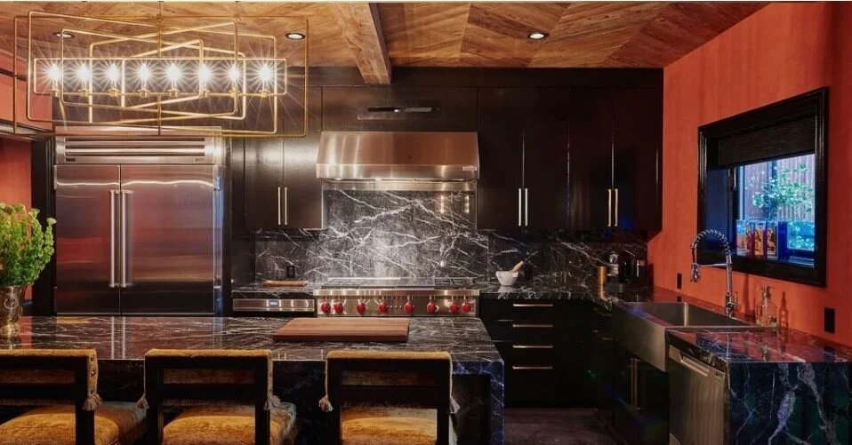 Modern kitchen with black marble countertops, dark cabinets, stainless steel appliances, a gold chandelier, and a red wall with a window.