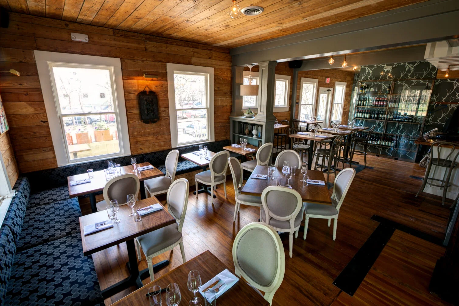 Interior of a cozy restaurant with wooden walls, large windows, and neatly set tables with glasses and napkins, featuring a mix of traditional and rustic decor.