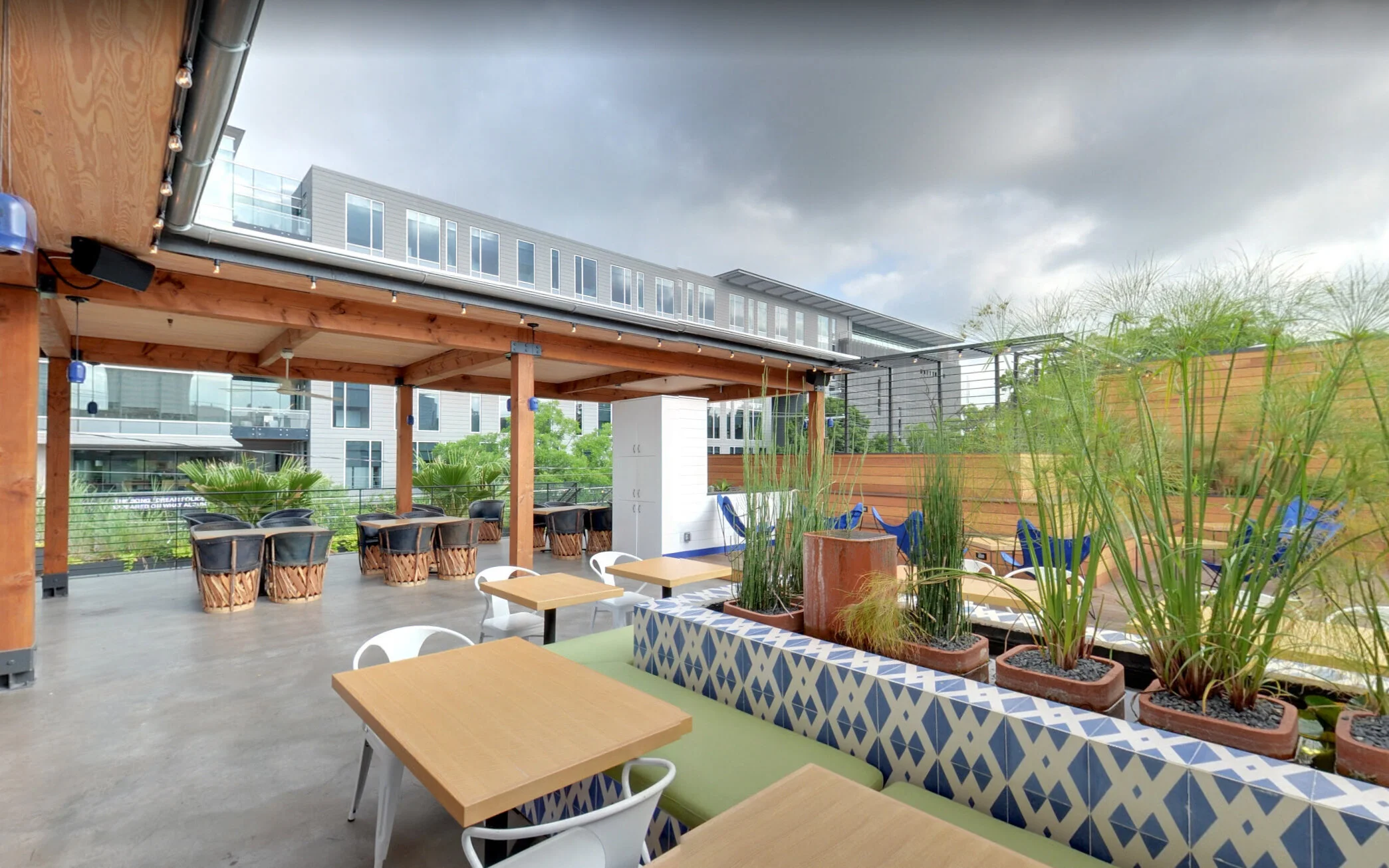 Rooftop patio with wooden beams, modern furniture, potted plants, and city buildings in the background under cloudy skies.