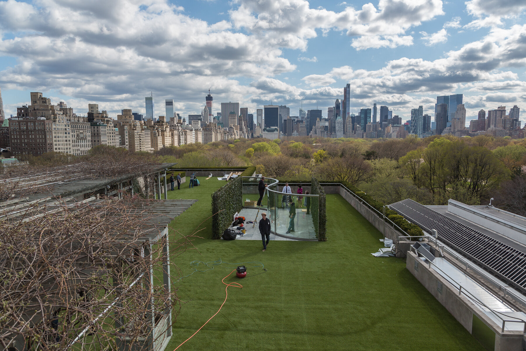 Rooftop garden with people, city skyline with tall buildings, trees, cloudy sky.